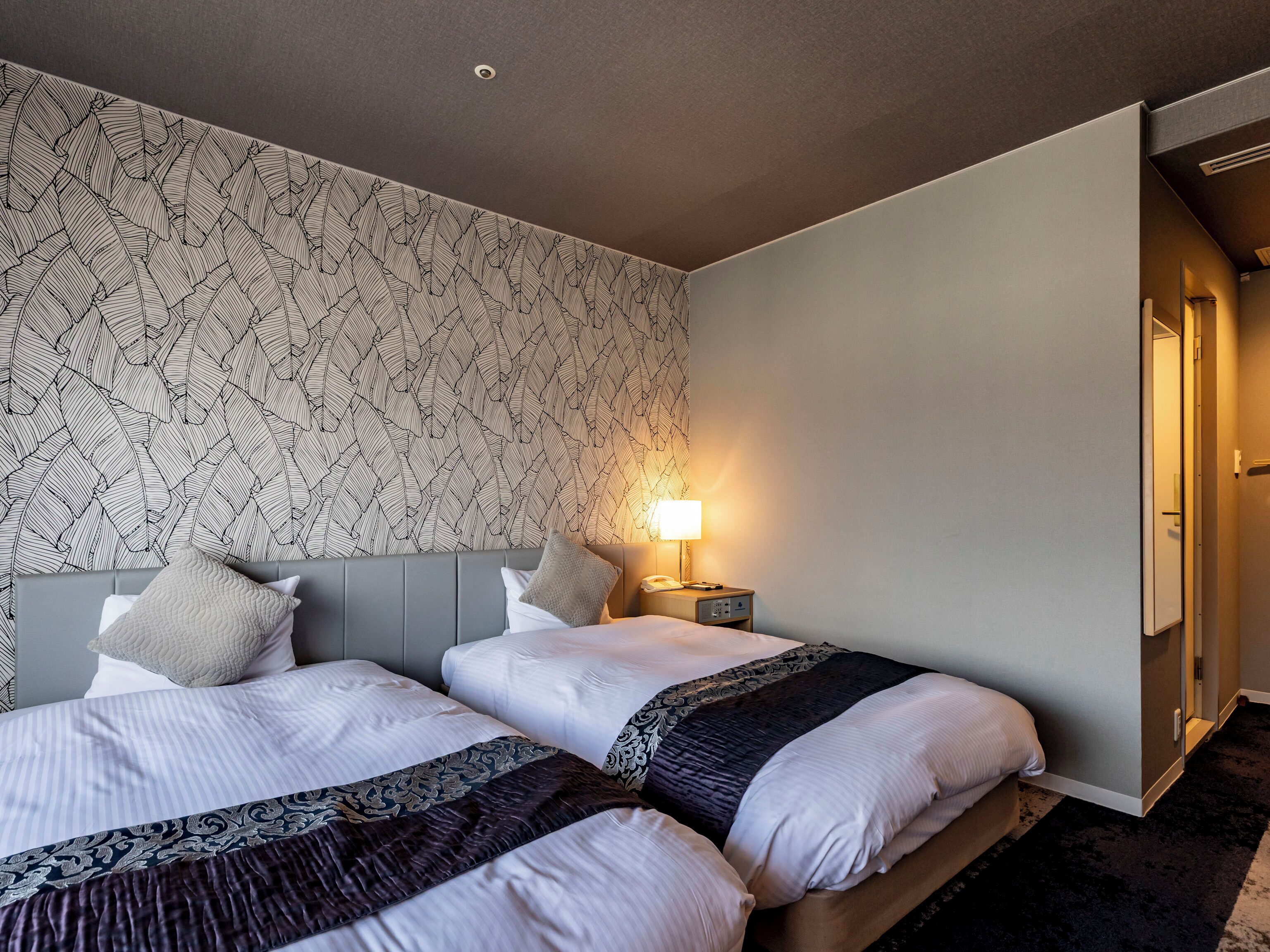 Une chambre d’hôtel avec deux lits, un papier peint à motifs, une table de chevet avec une lampe et une porte donnant sur une autre pièce.