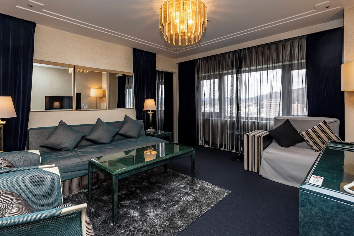 Ett modernt hotellrum med en mörkblå soffa, ett soffbord i glas, en randig fåtölj och ett stort fönster med gardiner.