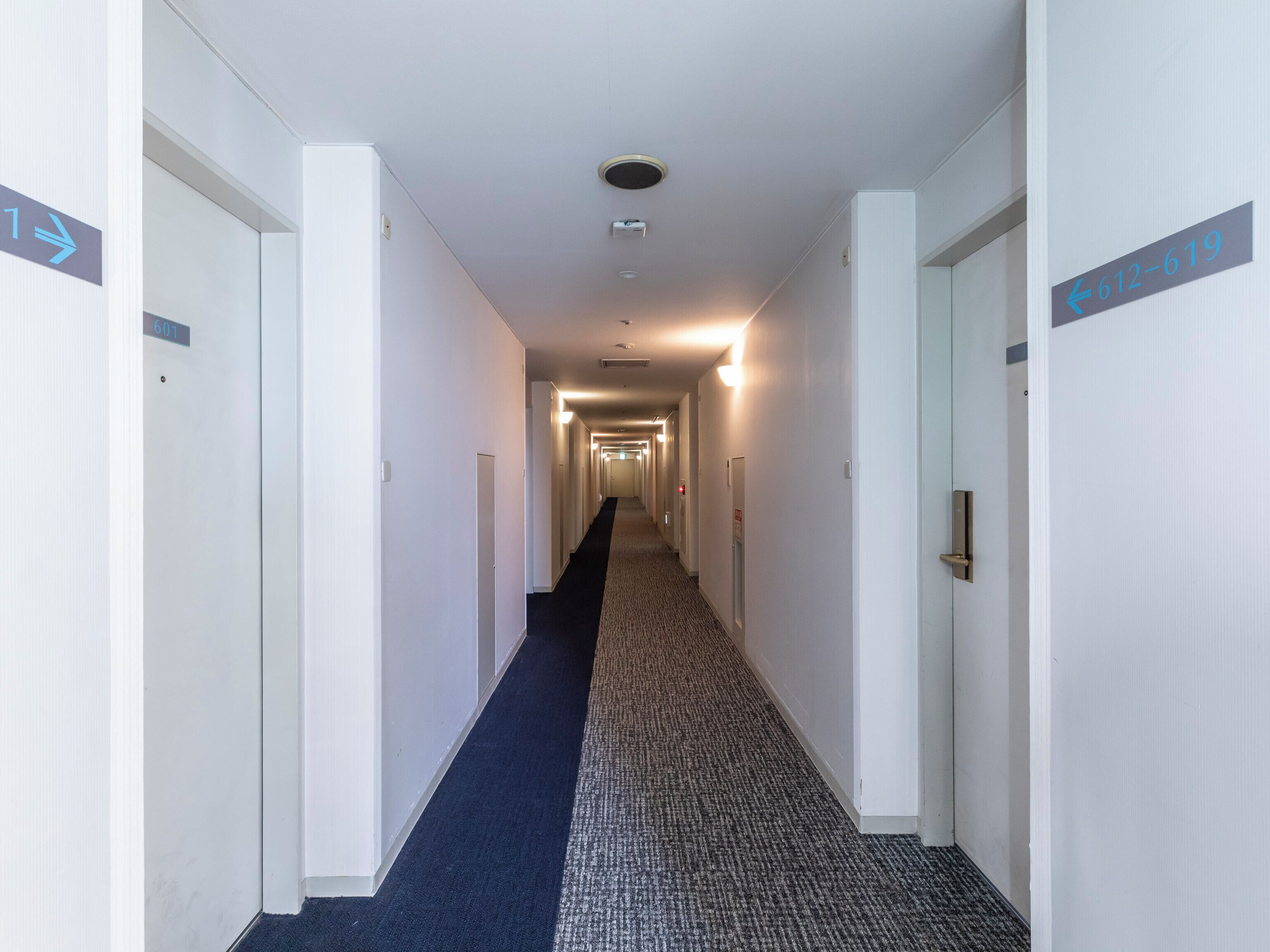 Un largo y estrecho pasillo de hotel con puertas a ambos lados, letreros con números y suelo alfombrado.