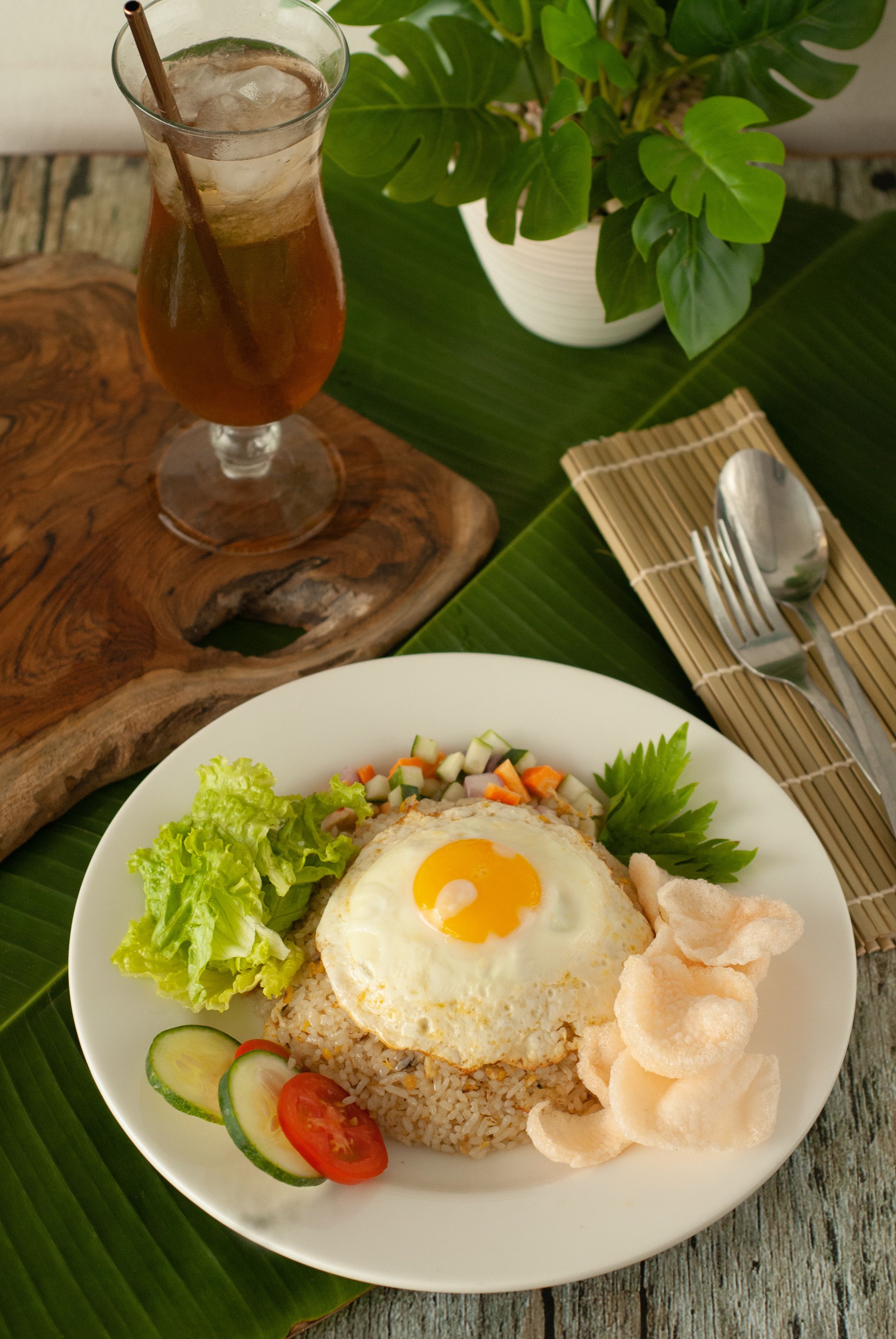 A plate of fried rice with vegetables and a fried egg, a glass of iced tea, and a plant in the background.