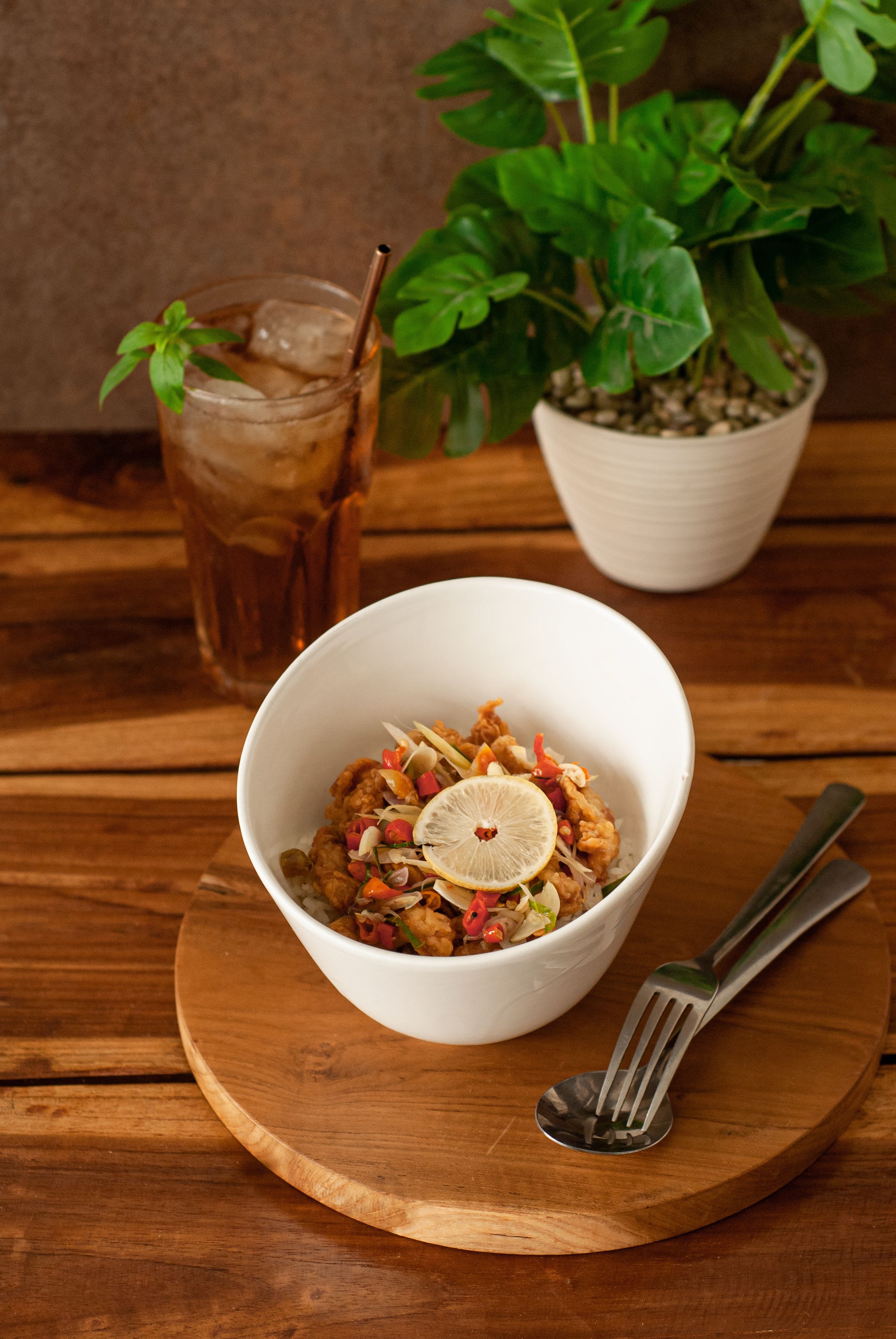 Uma tigela de comida sobre uma tábua de madeira, com um garfo ao lado, um copo de bebida gelada e uma planta em vasos ao fundo.