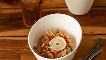 Uma tigela de comida sobre uma tábua de madeira, com um garfo ao lado, um copo de bebida gelada e uma planta em vasos ao fundo.
