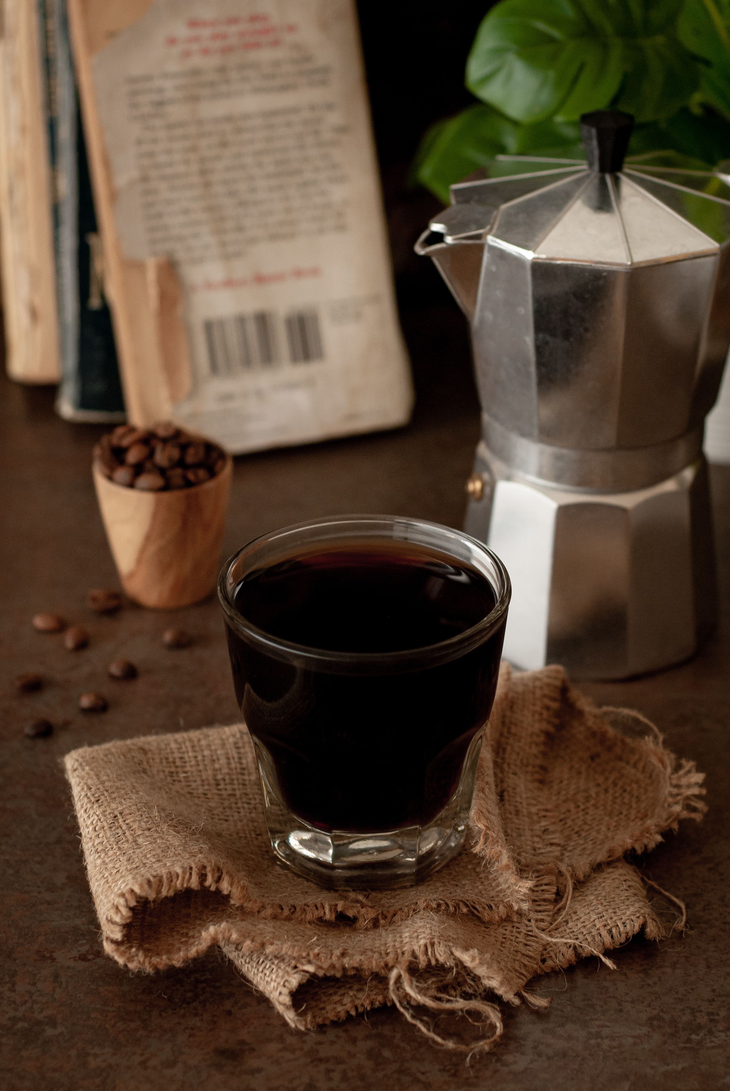 A cup of coffee on a burlap cloth, a coffee maker, and a wooden coffee cup filled with coffee beans.