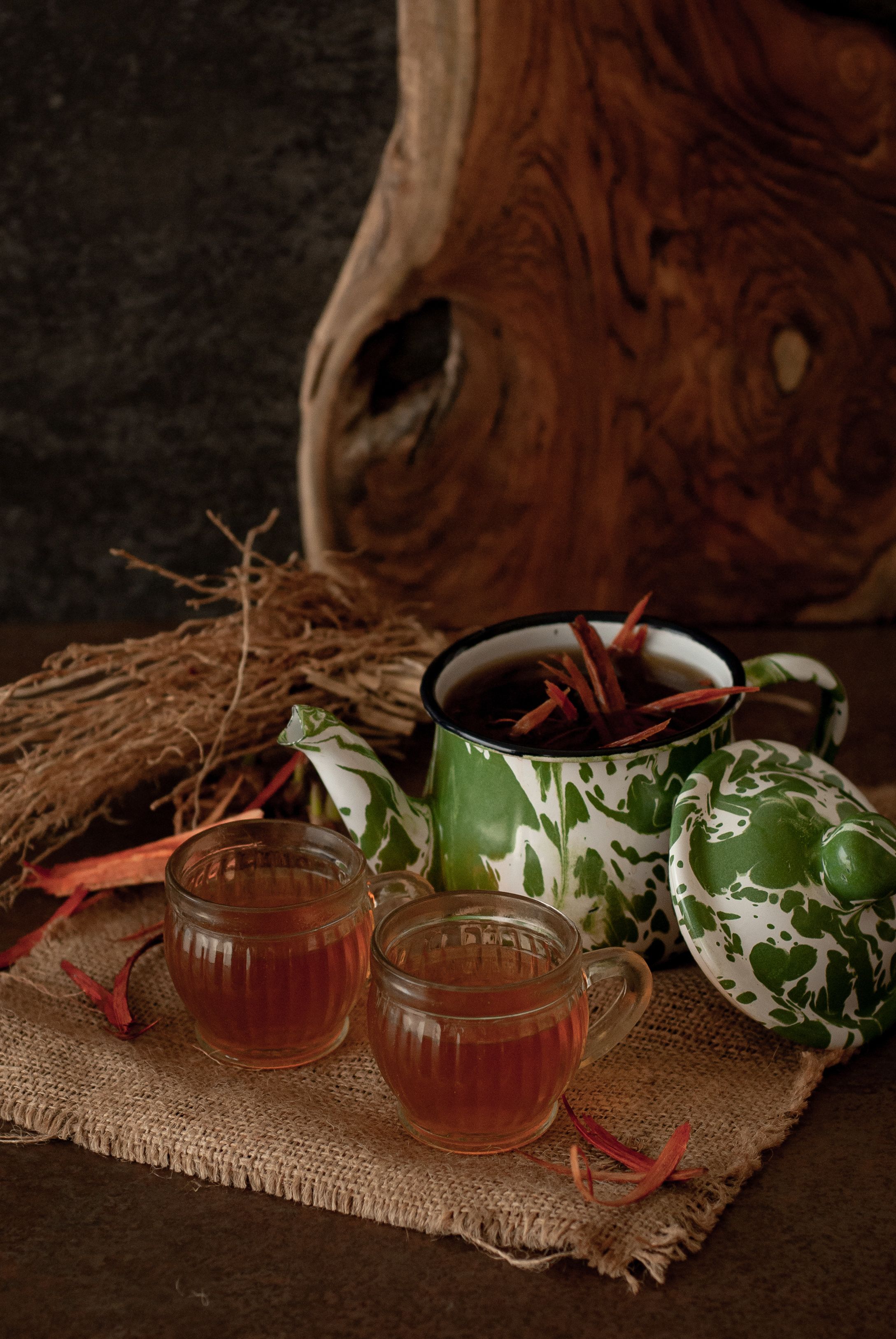 A rustic setting with a wooden table, a burlap cloth, a green and white patterned teapot, a green cup, and a glass mug filled with tea.