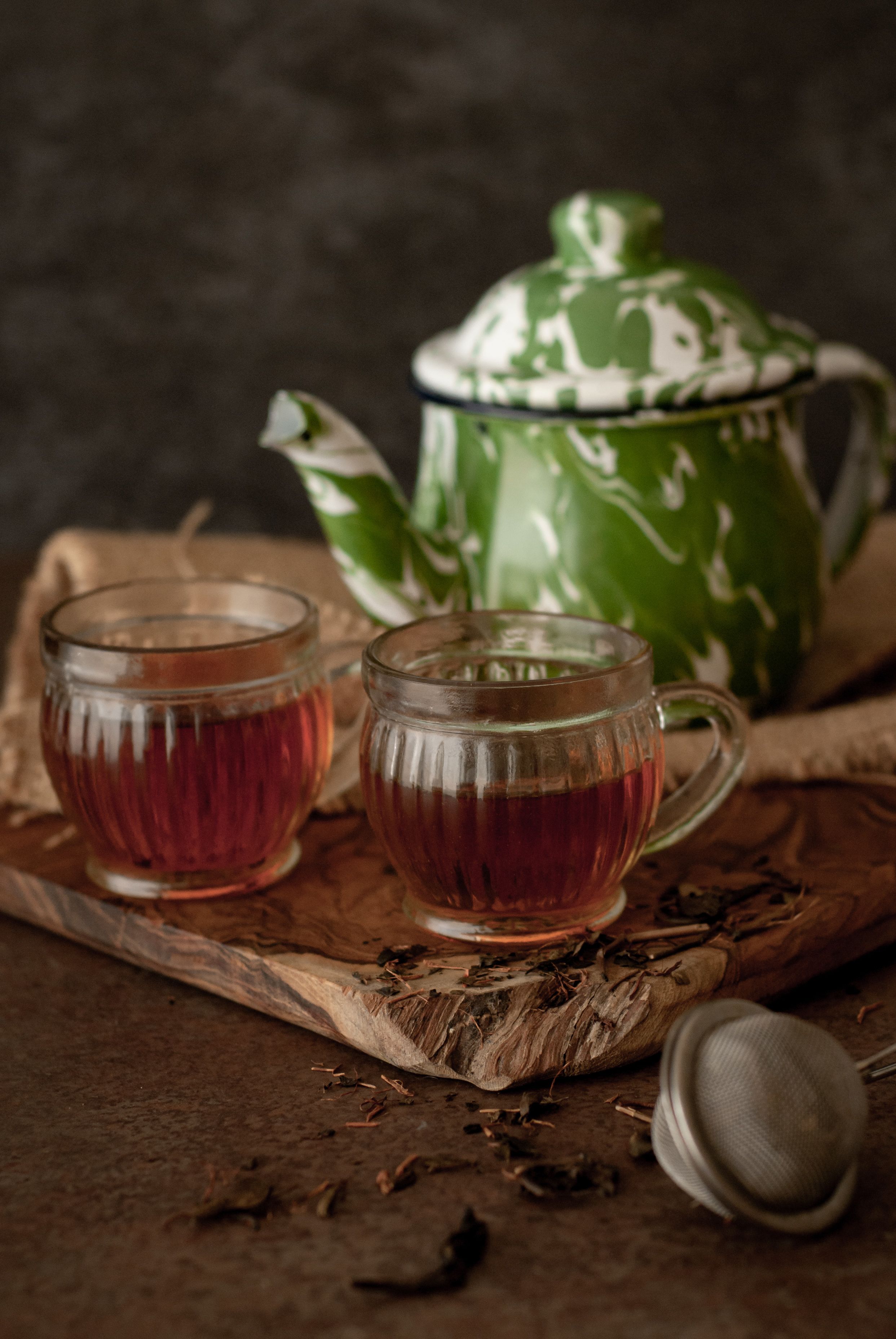 Dos tazas de vidrio transparentes con té sobre una bandeja de madera, junto con hojas de té sueltas, un colador de metal y una tetera verde y blanca con diseño.