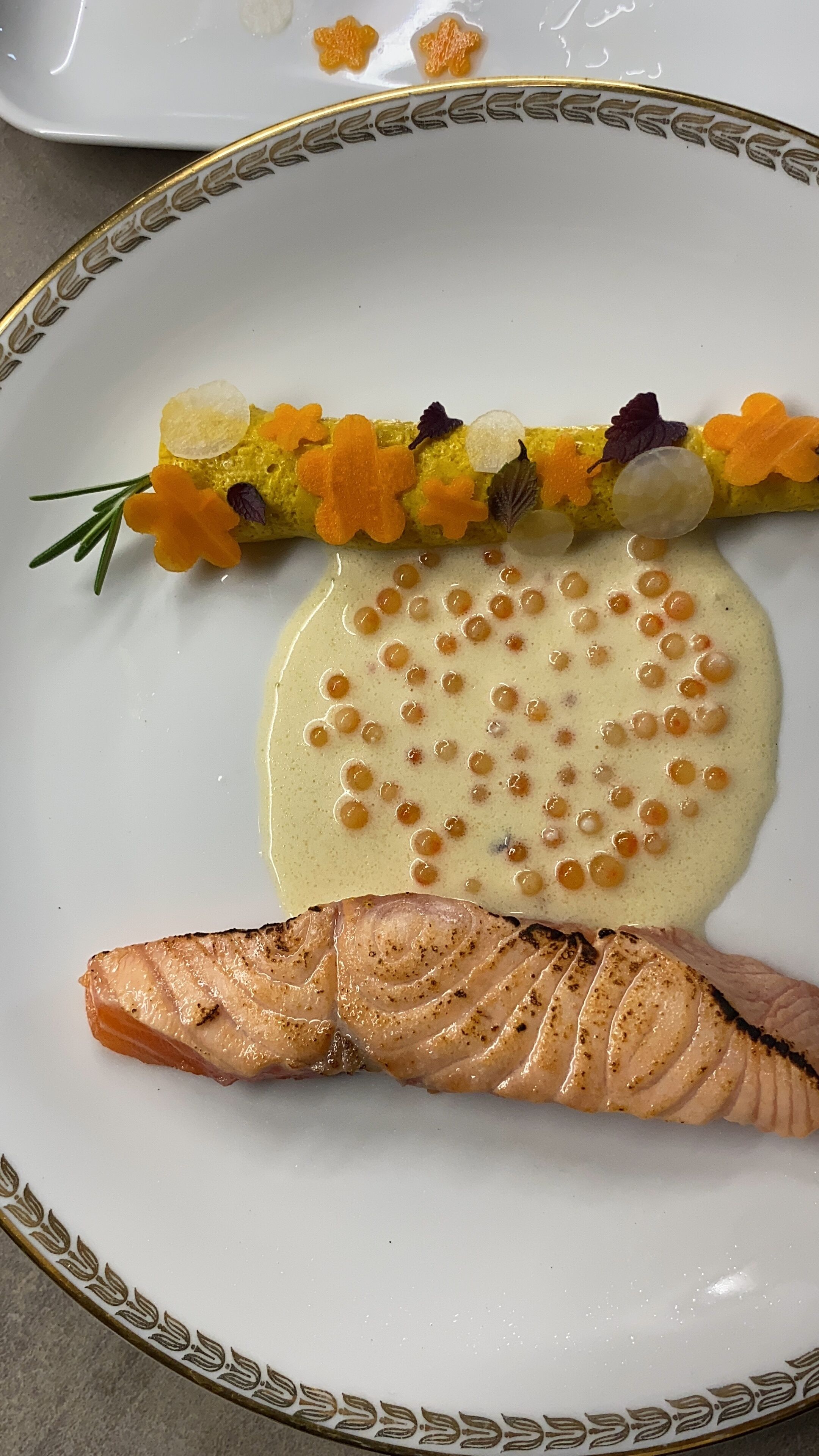 A plate with a salmon fillet, a decorative arrangement of orange and white elements, and a creamy sauce with small round items.