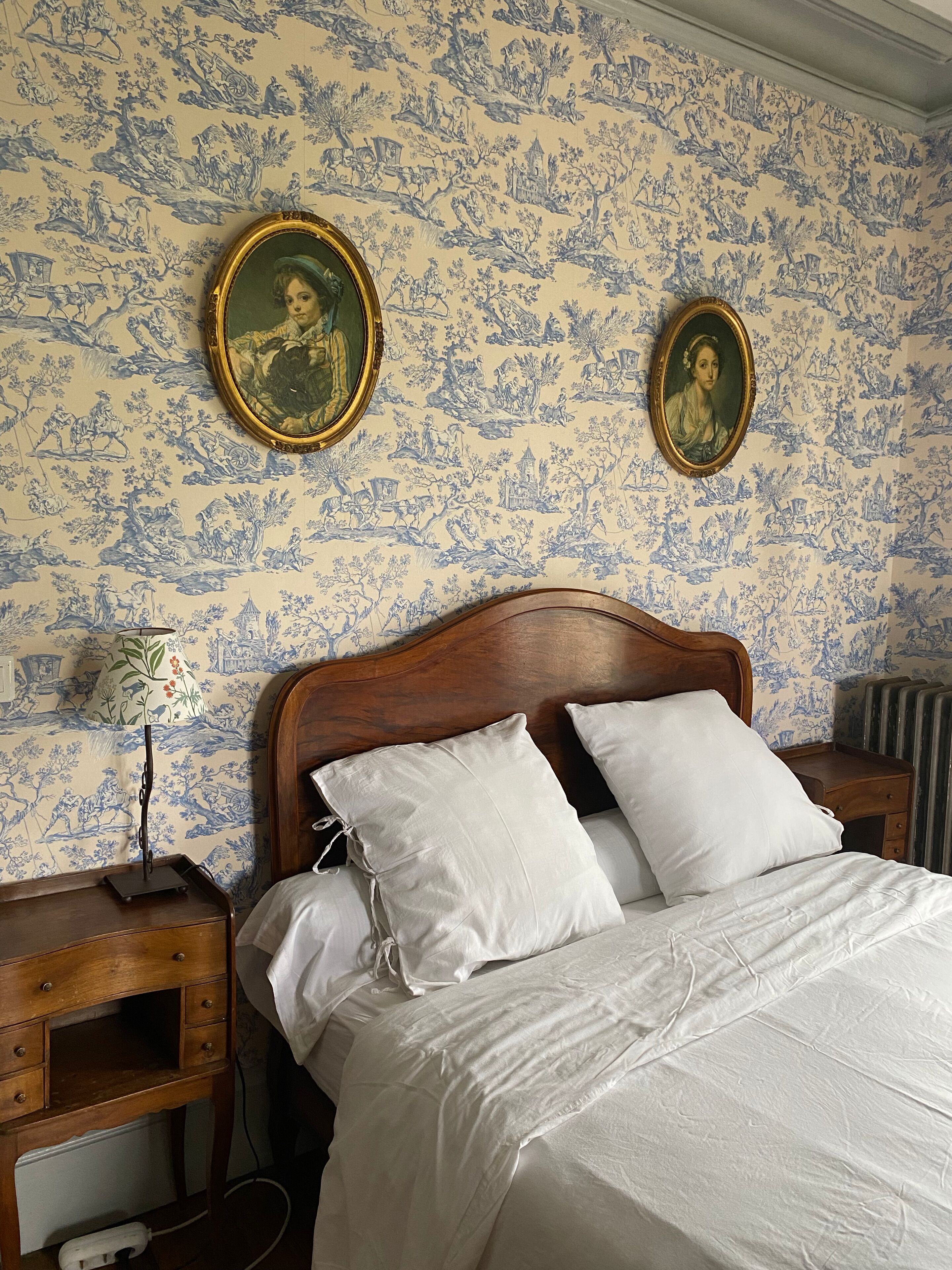 Un dormitorio con cabecera de madera, ropa de cama blanca, dos retratos enmarcados en la pared y una mesita de noche con lámpara.