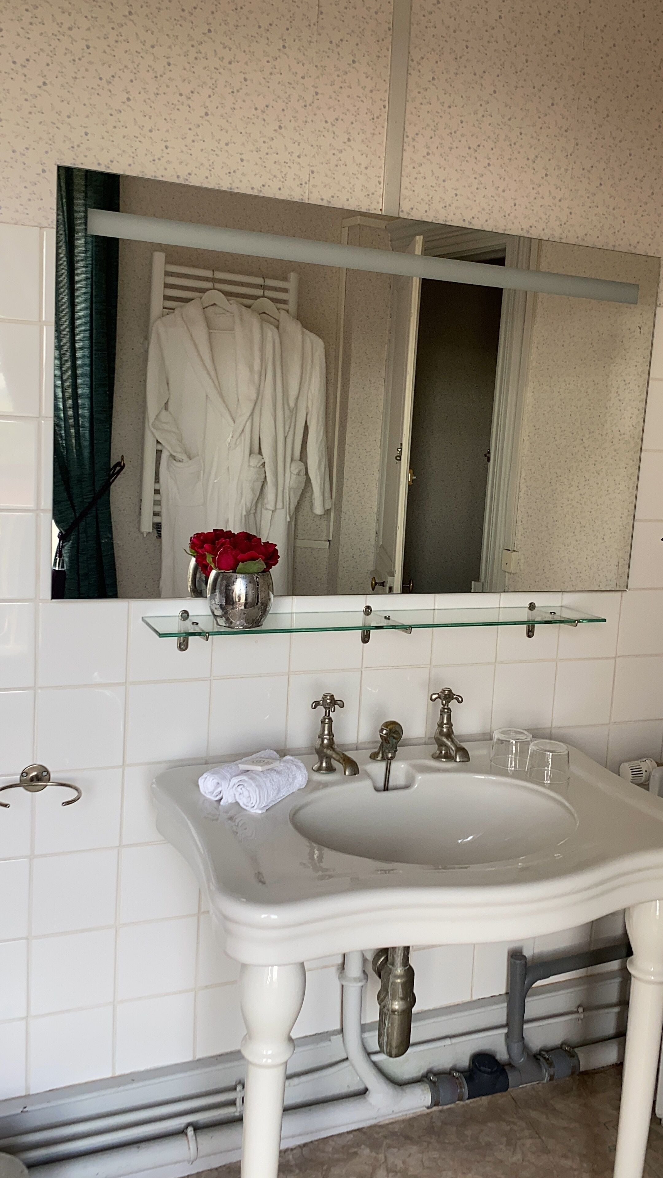 Ein Badezimmer mit einem weißen Waschbecken, einer Glasablage mit einer Vase roter Rosen und einem Spiegel über dem Waschbecken.