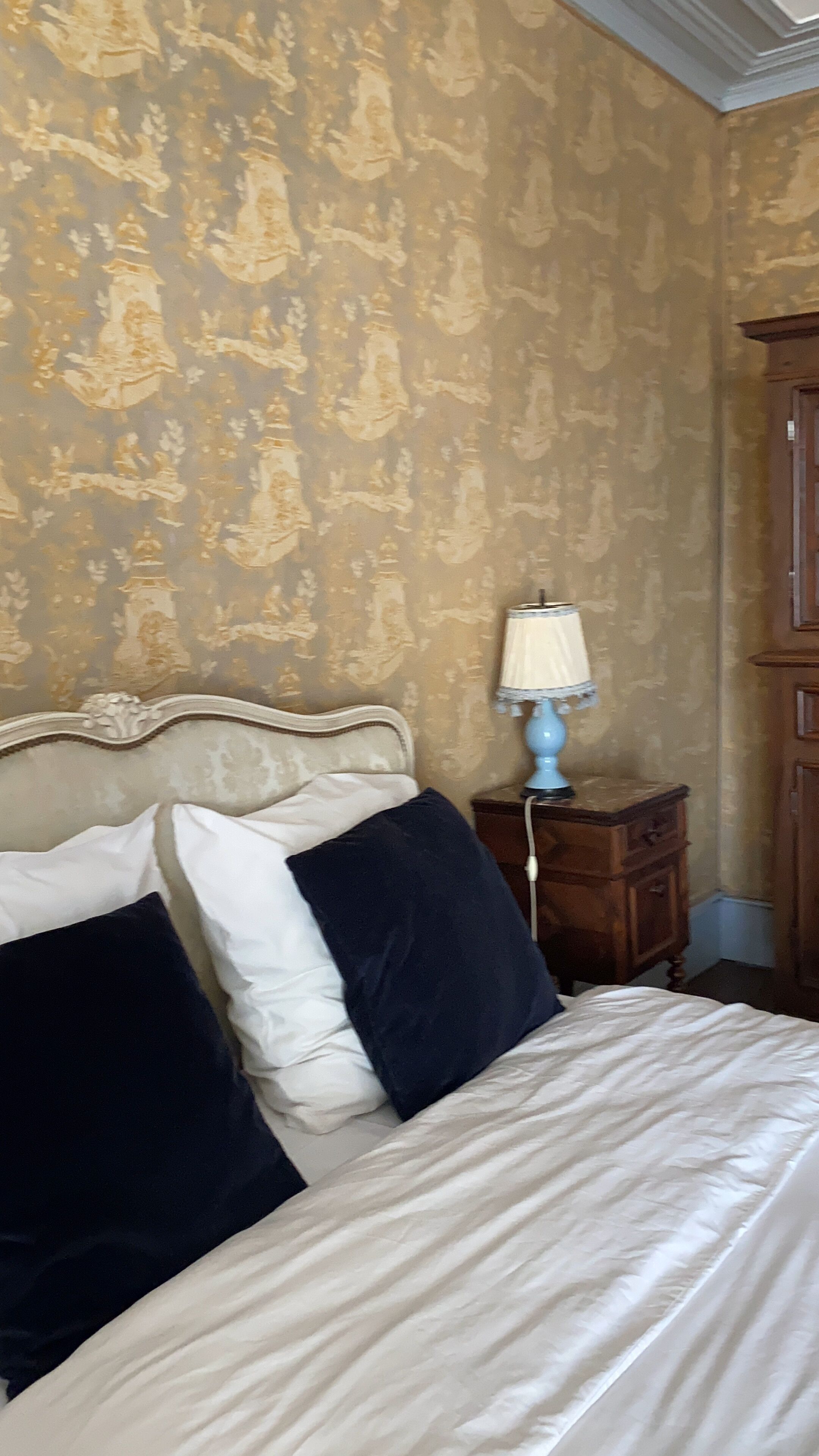 Una cama con almohadas blancas y azul marino, una lámpara azul sobre una mesita de noche de madera, y un armario de madera.