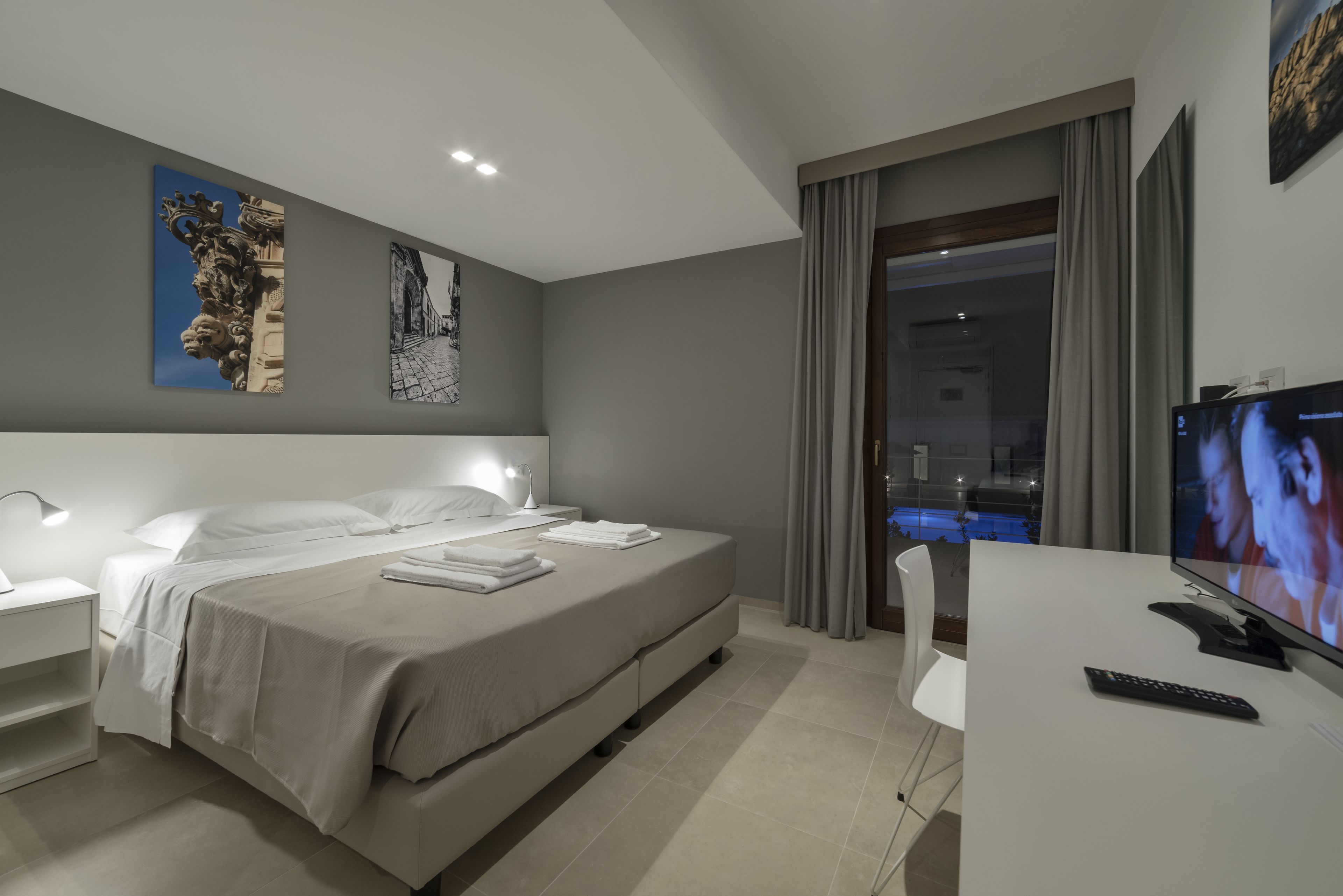 A modern hotel room with a large bed, bedside tables, a desk with a TV, and a balcony with a pool view.