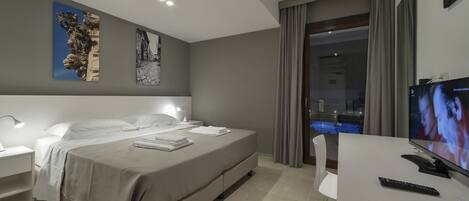 A modern hotel room with a large bed, bedside tables, a desk with a TV, and a balcony with a pool view.