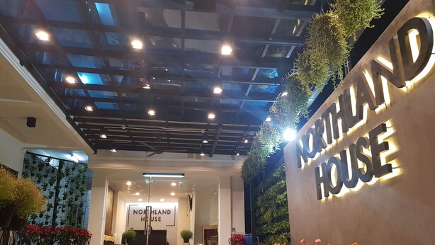 Une entrée moderne d’un immeuble, avec une enseigne indiquant « NORTHLAND HOUSE », et un espace paysager bien entretenu, agrémenté de plantes en pot et de fleurs.