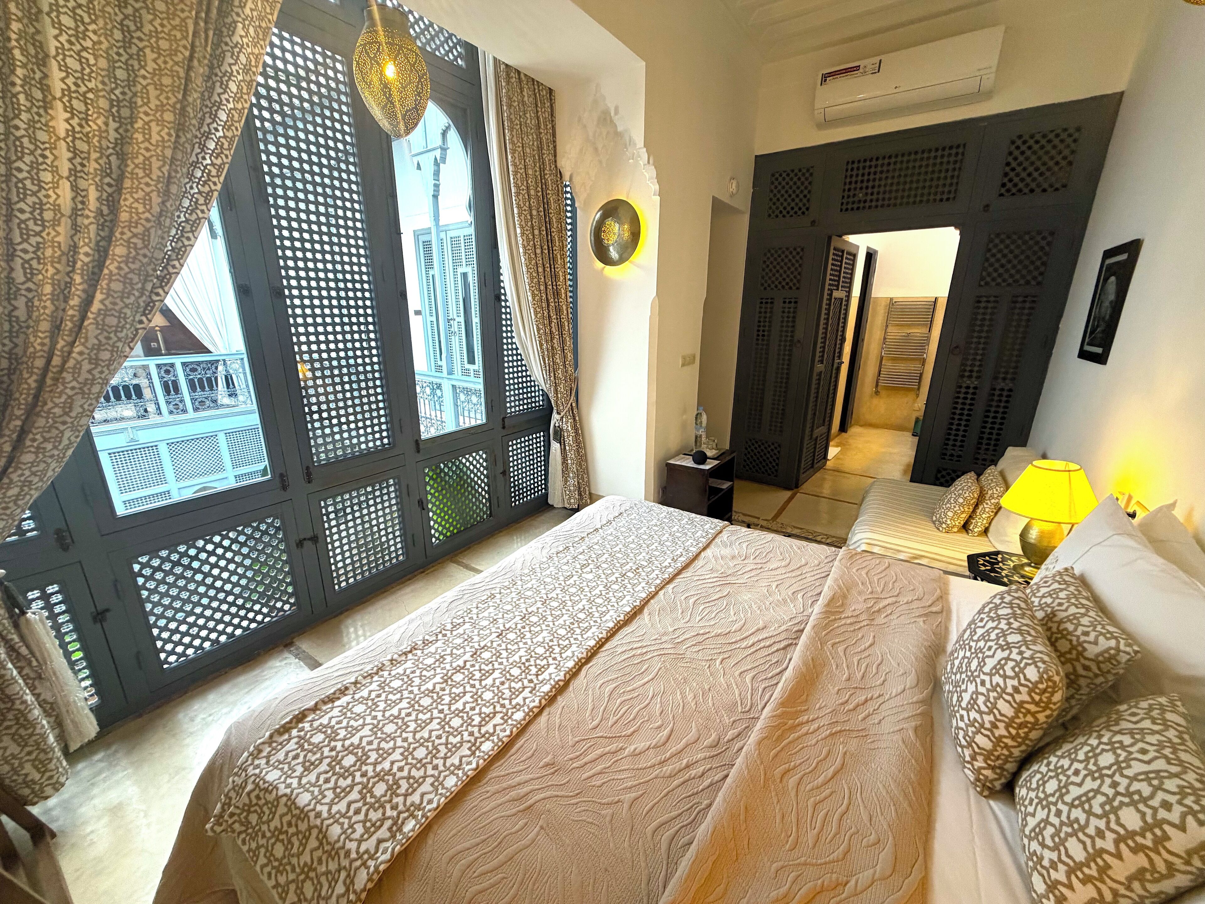 A bedroom with a bed, patterned bedding, a nightstand, a lamp, and a view through a window with a lattice design.