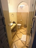 Un baño con inodoro, lavamanos y zona de ducha, con suelo de baldosas y una toalla en el piso.