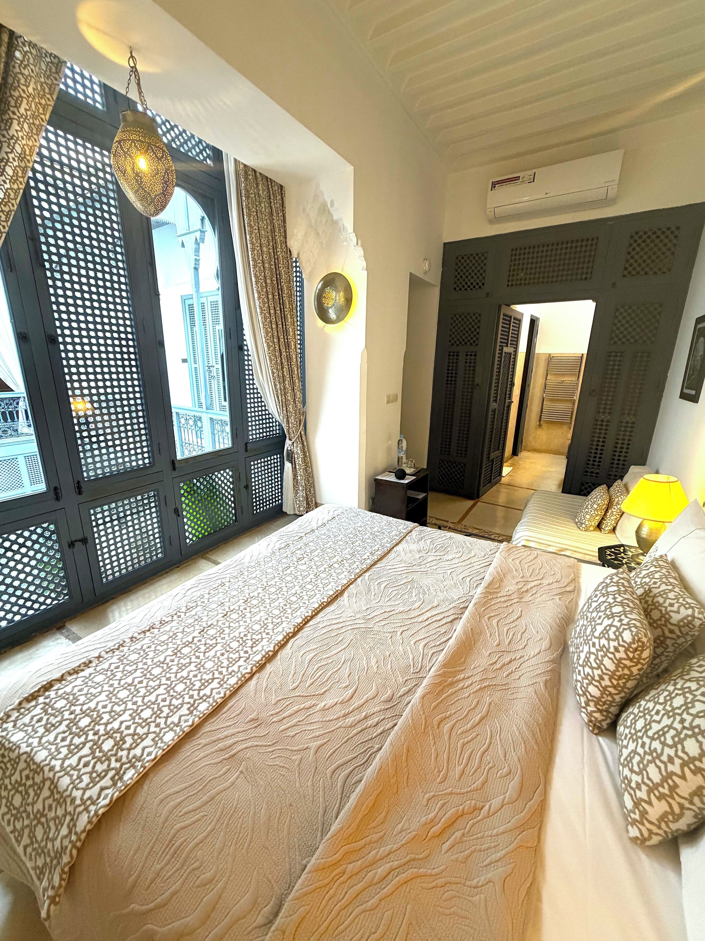 A bedroom with a bed, a hanging light fixture, a window with patterned curtains, and an air conditioning unit.