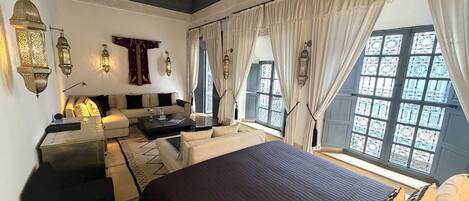 A room with a bed, sofa, and coffee table, featuring ornate wall sconces and a view of the outside.