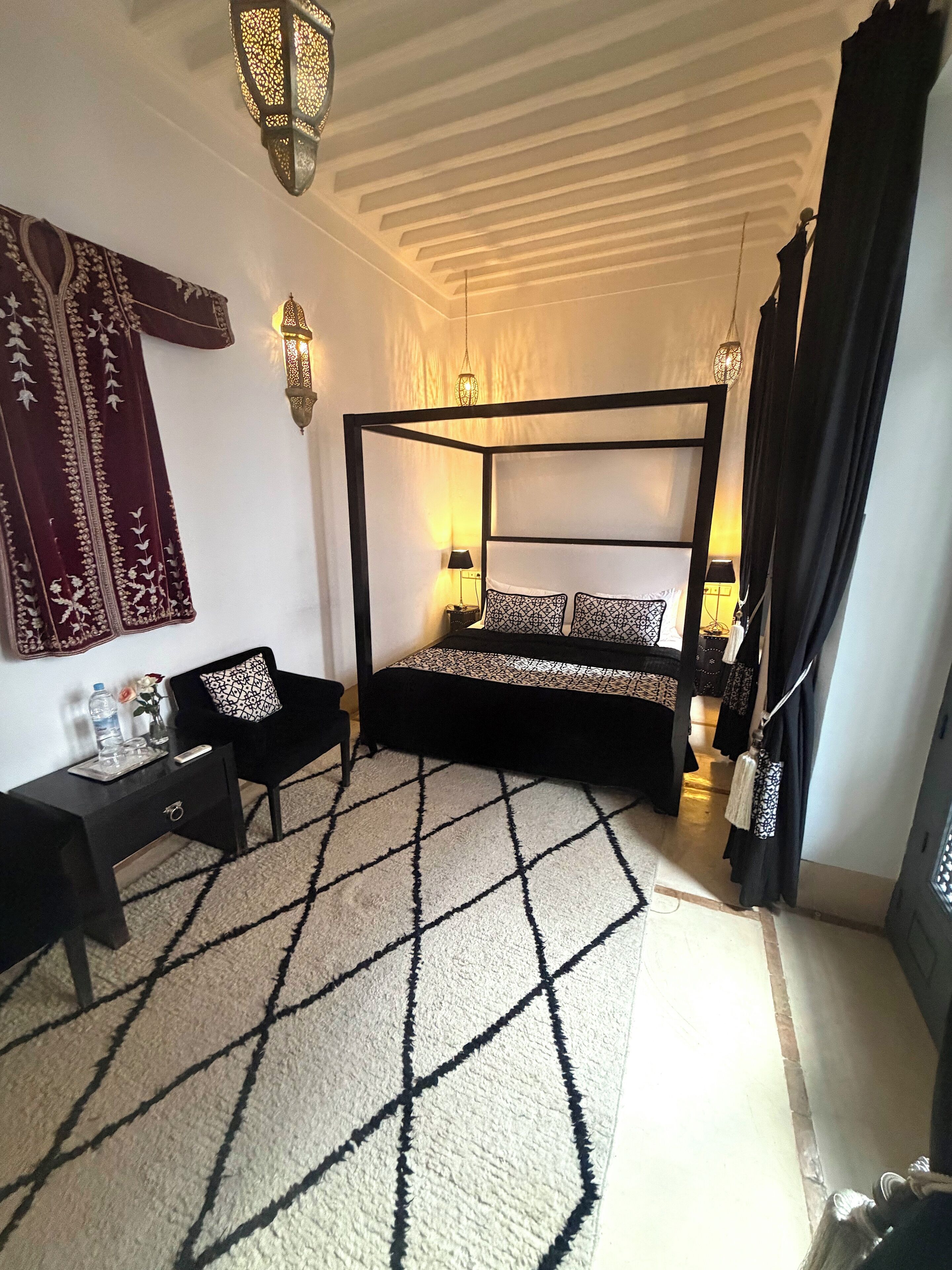 A bedroom with a four-poster bed, a nightstand with a lamp, a chair, and a decorative tapestry.