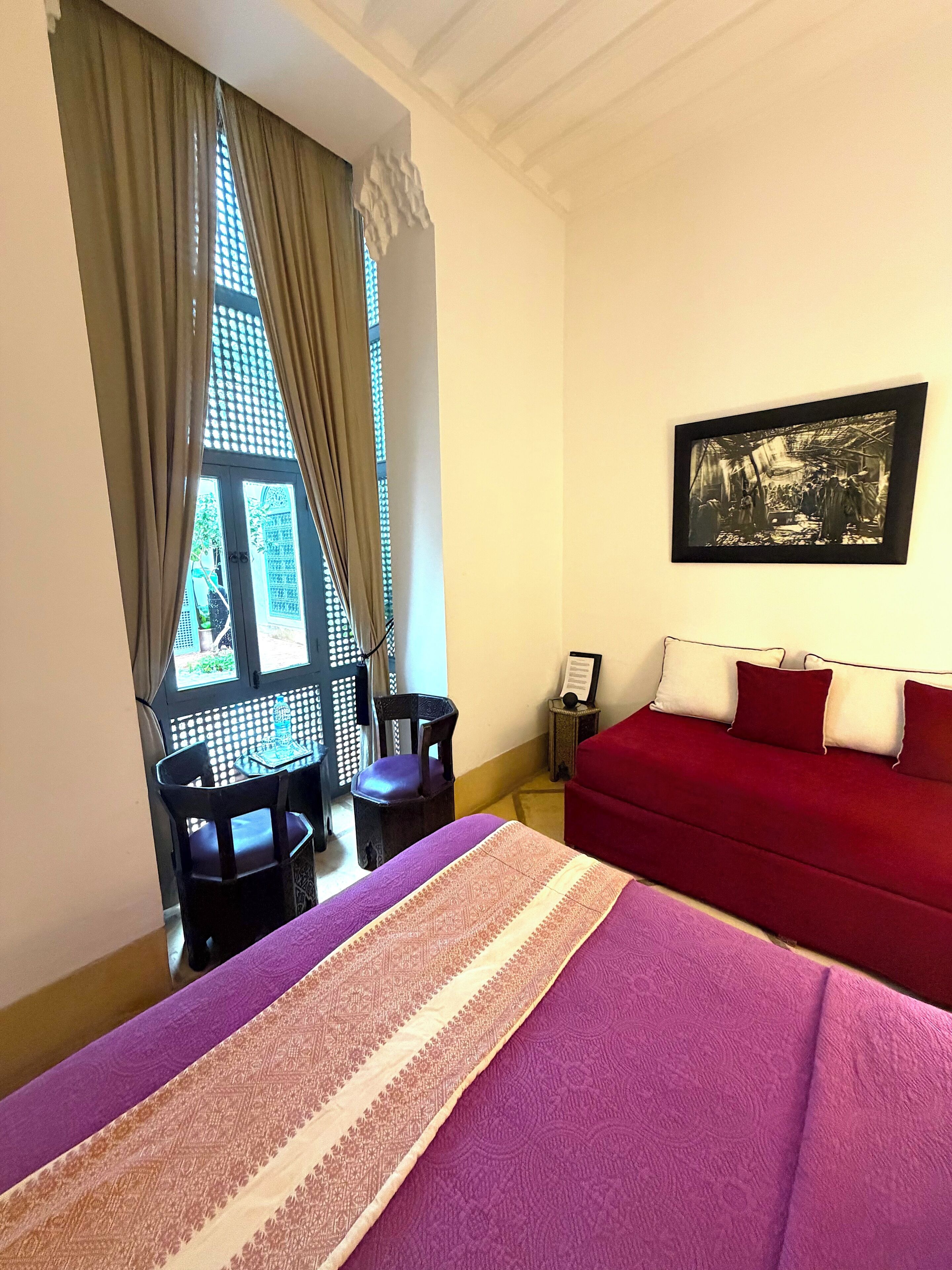 Una habitación de hotel con una cama, un sofá rojo, un cuadro en la pared, una ventana con cortinas y una mesa con sillas.