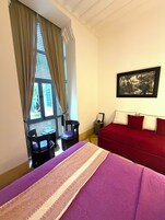 A hotel room with a bed, a red sofa, a framed picture on the wall, a window with curtains, and a table with chairs.