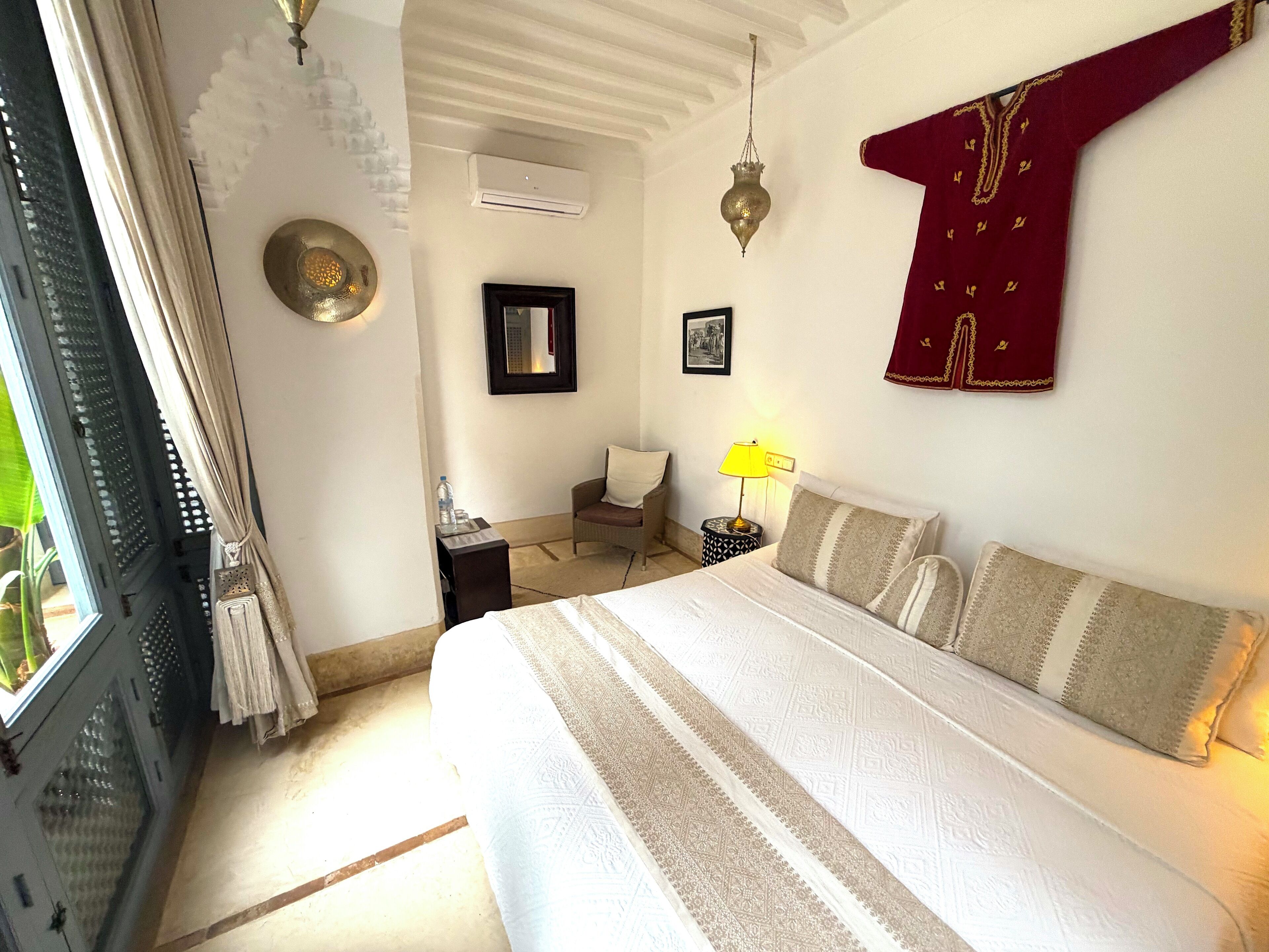 Un dormitorio con cama, mesita de noche, lámpara, silla, ventana con cortinas y aire acondicionado de pared.