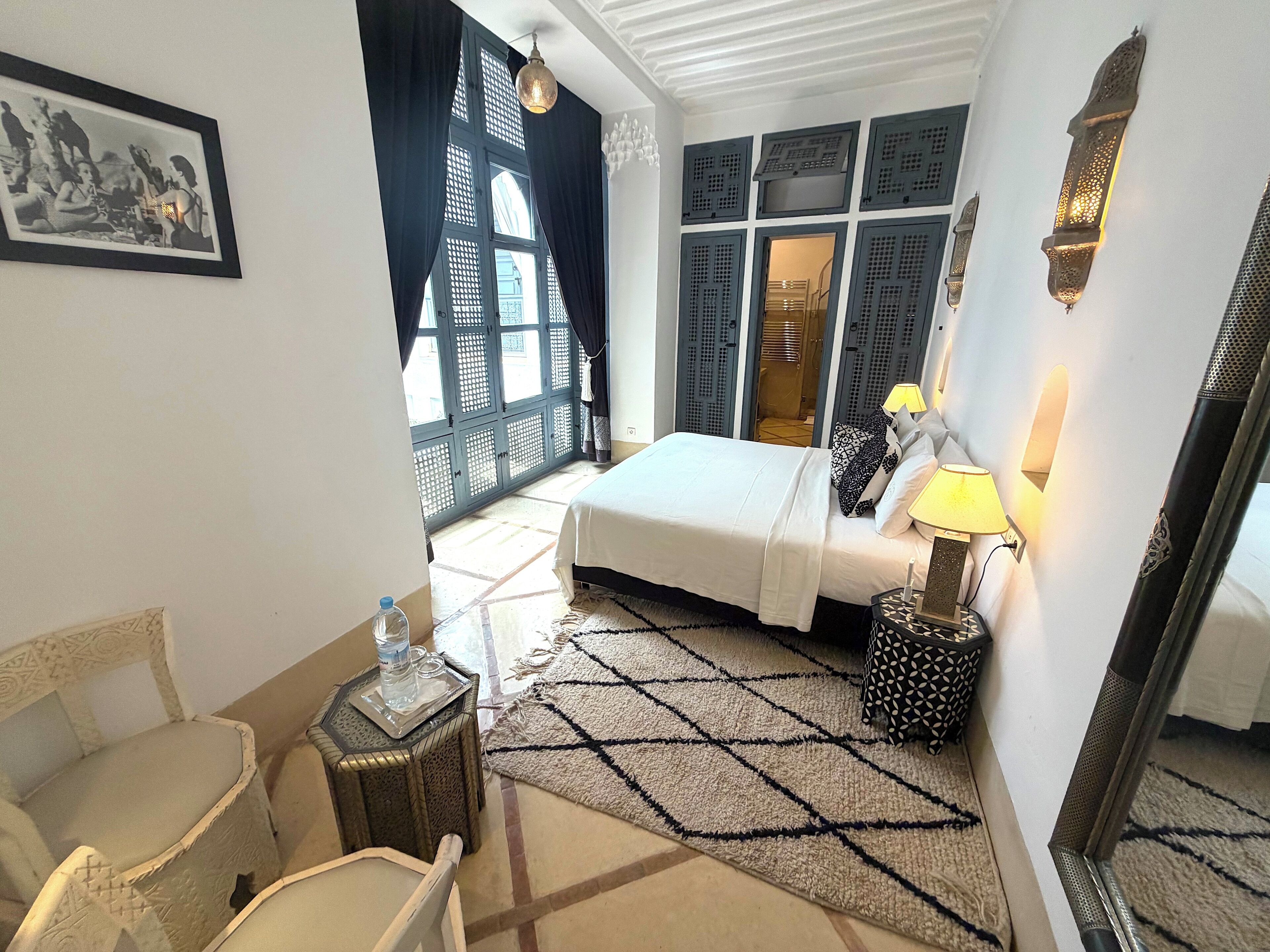 A hotel room with a bed, a side table with a lamp, a chair, a patterned rug, and a large window with curtains.