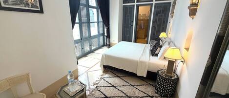 A hotel room with a bed, a side table with a lamp, a chair, a patterned rug, and a large window with curtains.