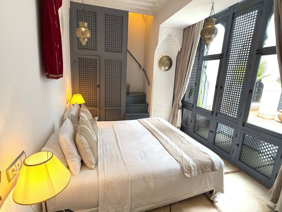 Un dormitorio con cama, mesita de noche, lámpara, un atuendo rojo en la pared, una escalera y una vista a través de una ventana con celosía.