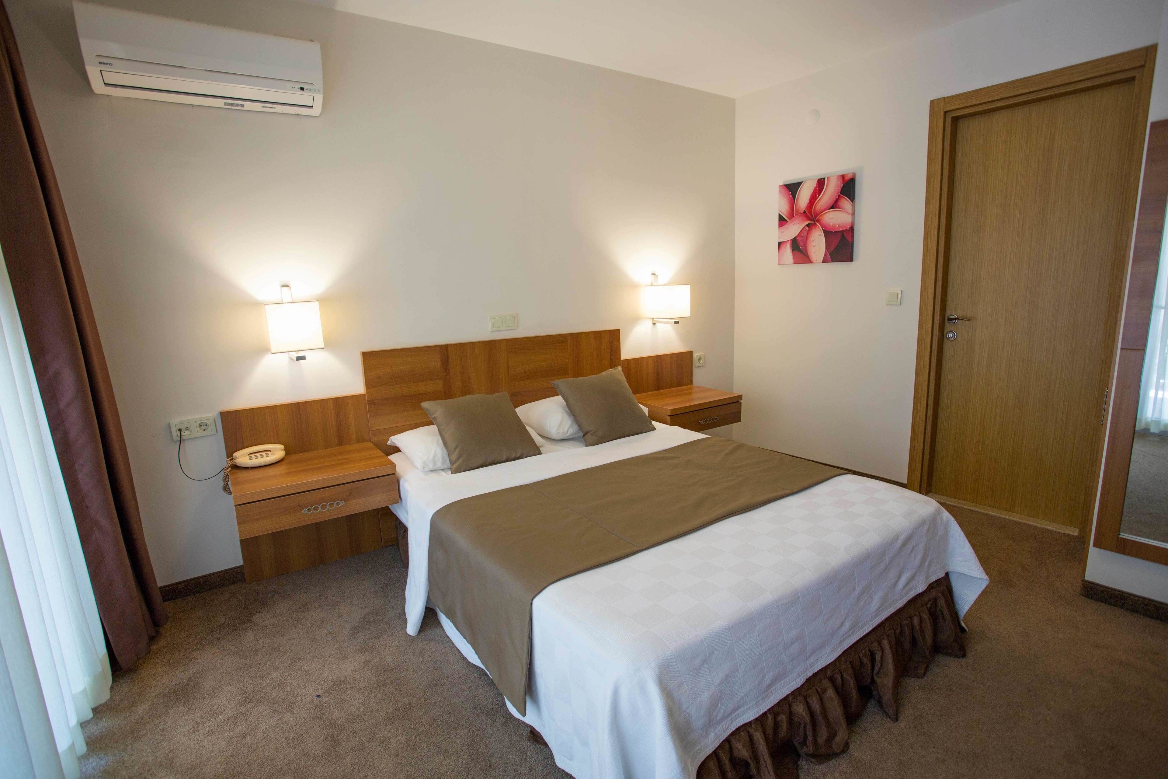 A hotel room with a bed, bedside tables, an air conditioner, a nightstand, a lamp, and a door.