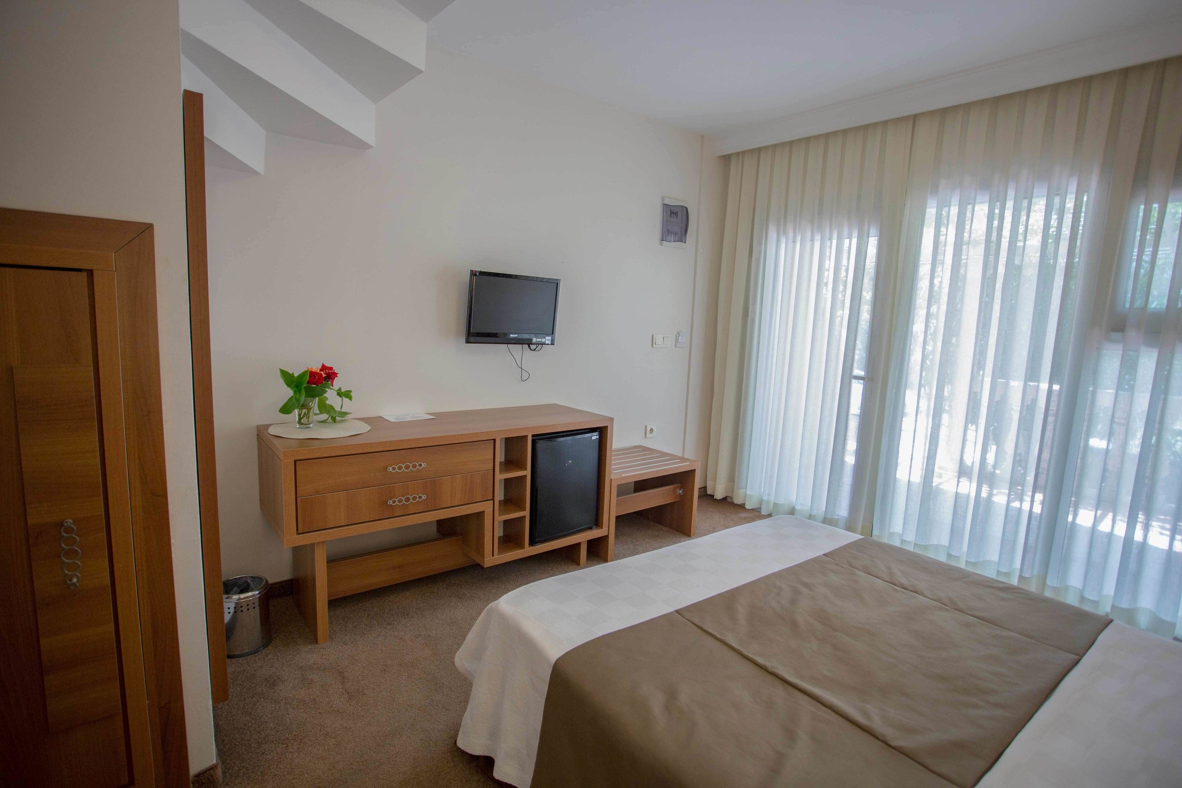 Habitación de hotel con cama, escritorio de madera, televisión, refrigerador pequeño y ventana con cortinas.