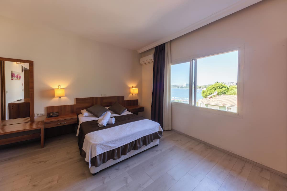 Habitación de hotel con cama, mesitas de noche, un espejo y un amplio ventanal con vistas al mar.