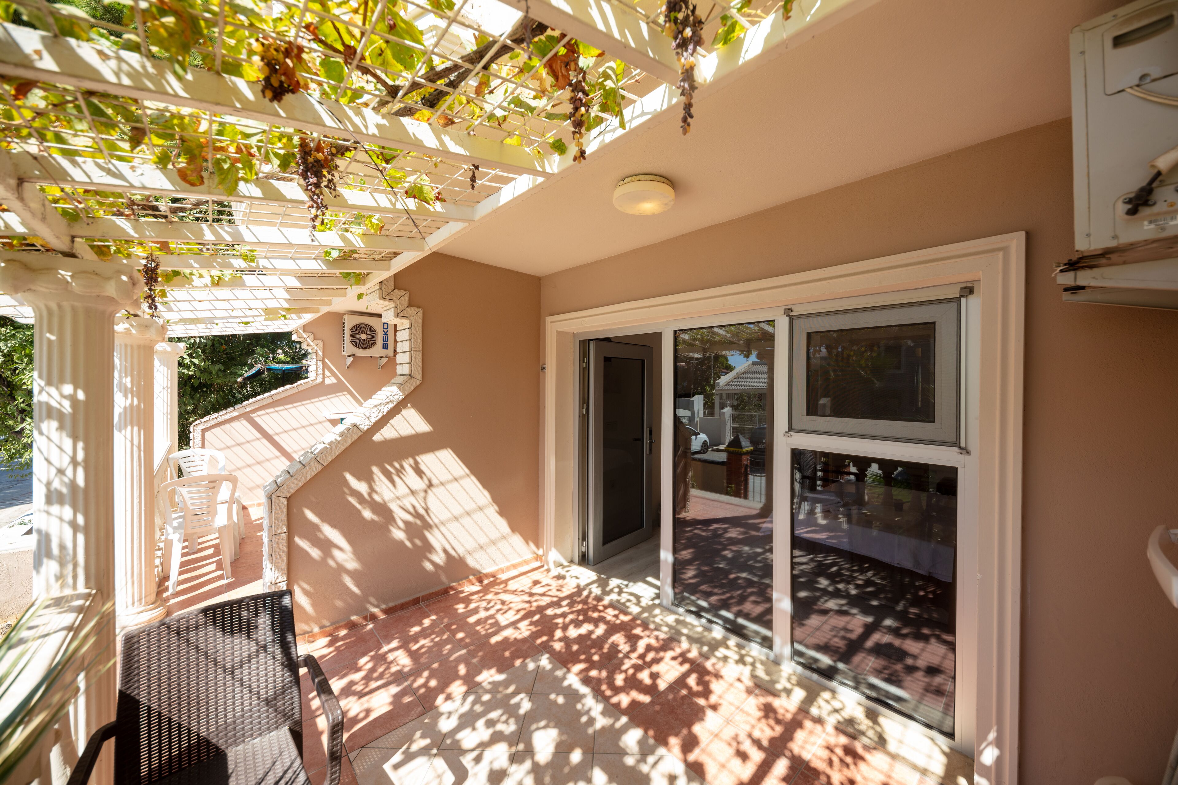 Un patio cubierto con pérgola, mobiliario exterior y un ventanal corredera que da a un espacio techado.