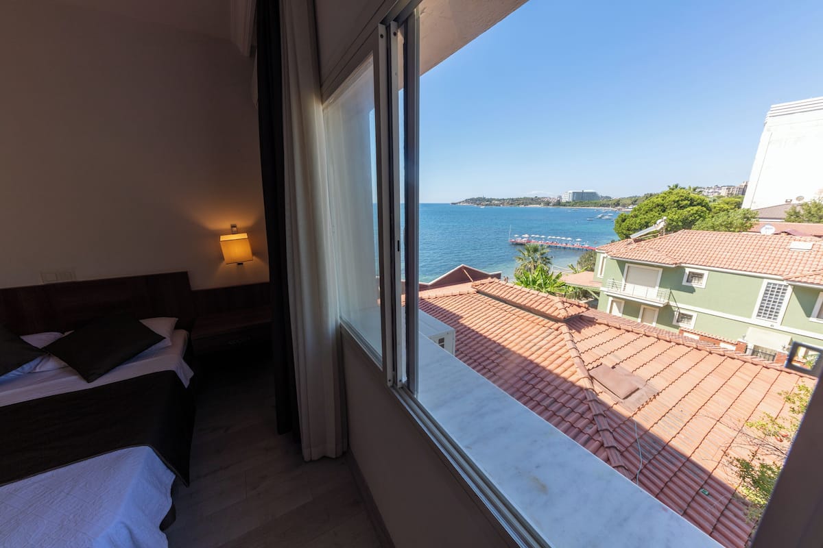 Un cuarto de hotel con cama, mesita de noche y ventana con vistas al mar y edificios.