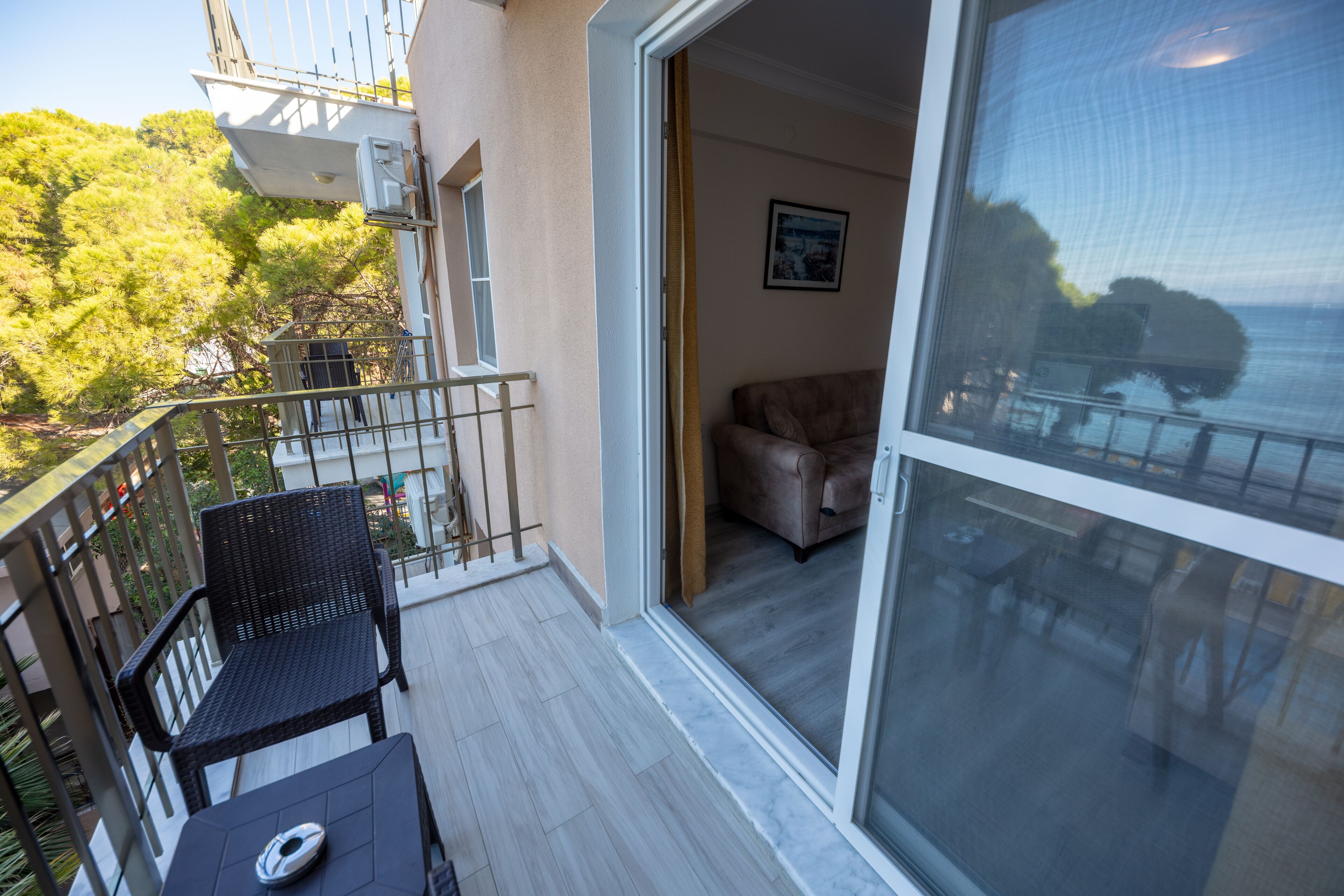 A balcony with a table and chairs, a sliding glass door leading to an interior room with a sofa, and a view of trees and a building.