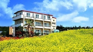 A multi-story hotel with balconies, located on a hill with a yellow flower field in the foreground. - Memory Tour (Seogwipo)