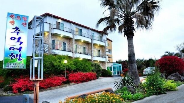 A multi-story hotel with balconies, surrounded by landscaped gardens with red and yellow flowers, and a palm tree. - Memory Tour (Seogwipo)