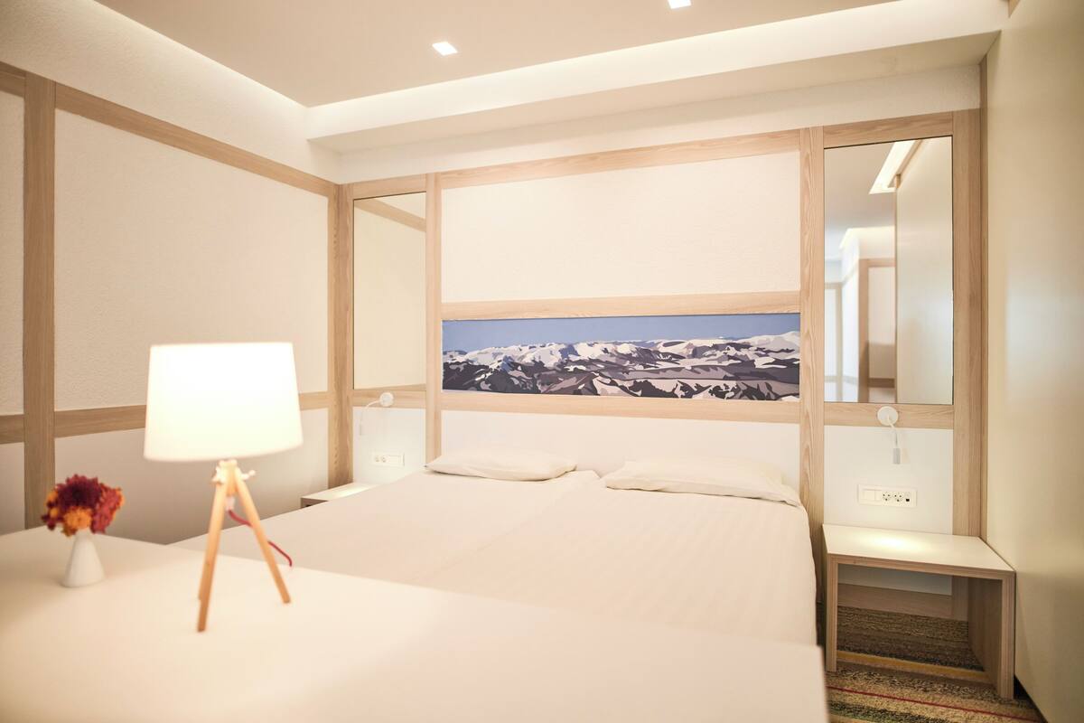 A hotel room with a large bed, a nightstand, a lamp, and a painting of a mountain landscape.