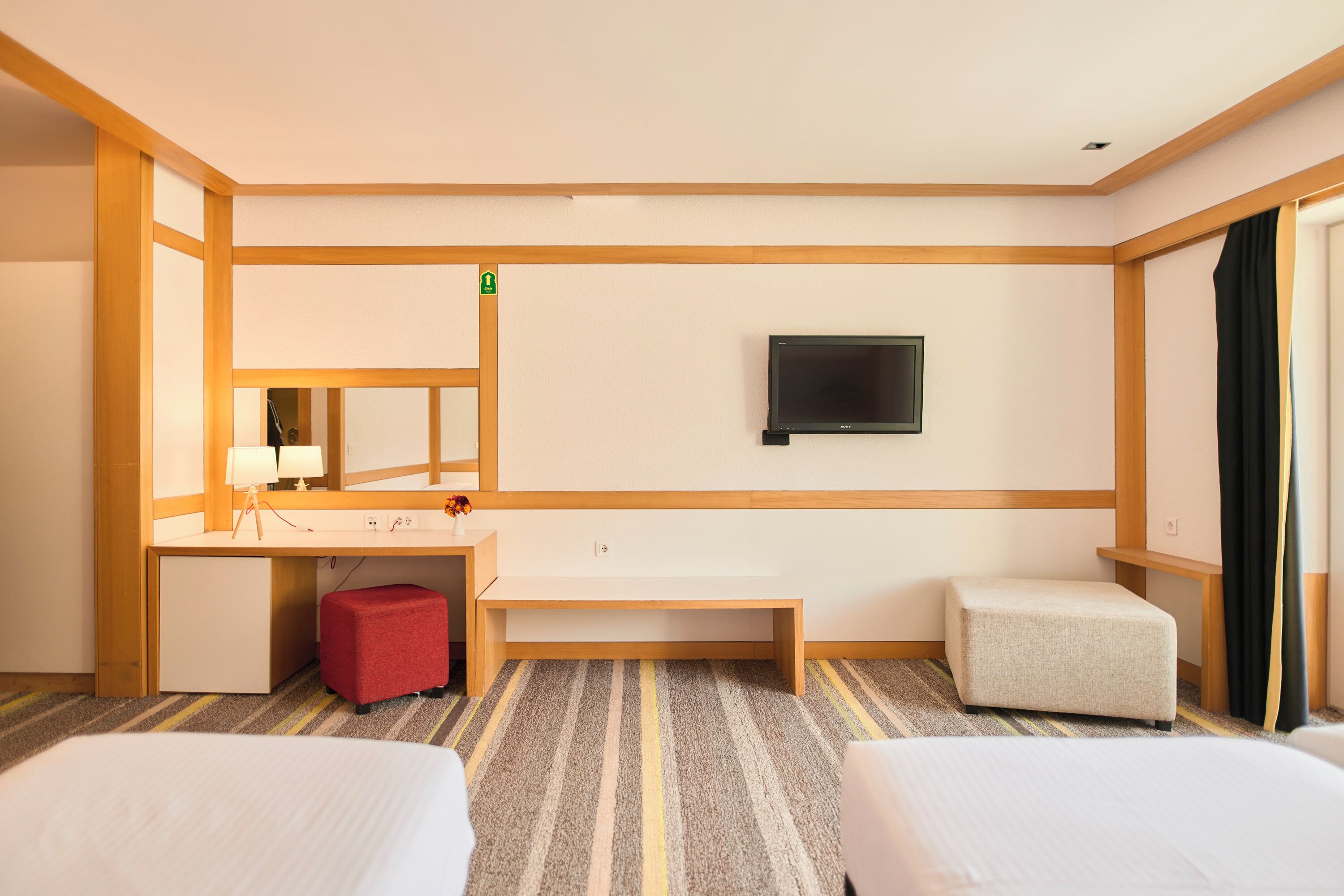 Quarto de hotel com duas camas, uma escrivaninha, televisão e um banquinho vermelho pequeno.