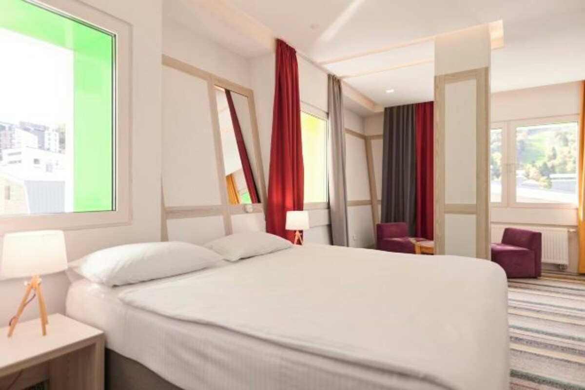 Quarto de hotel com uma cama grande, uma mesa de cabeceira com abajur, uma cadeira e vista para a cidade pela janela.