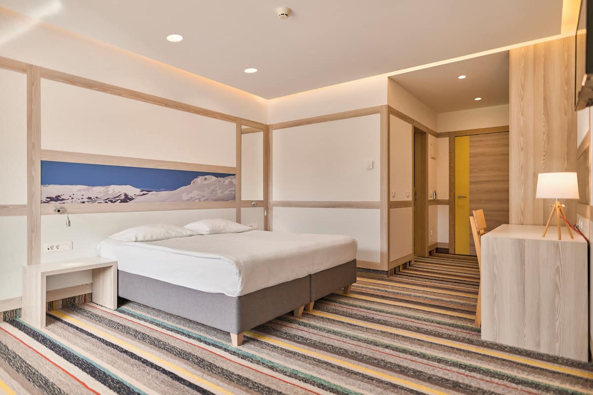 A modern hotel room with a large bed, a desk, a chair, and a view of a snowy mountain.