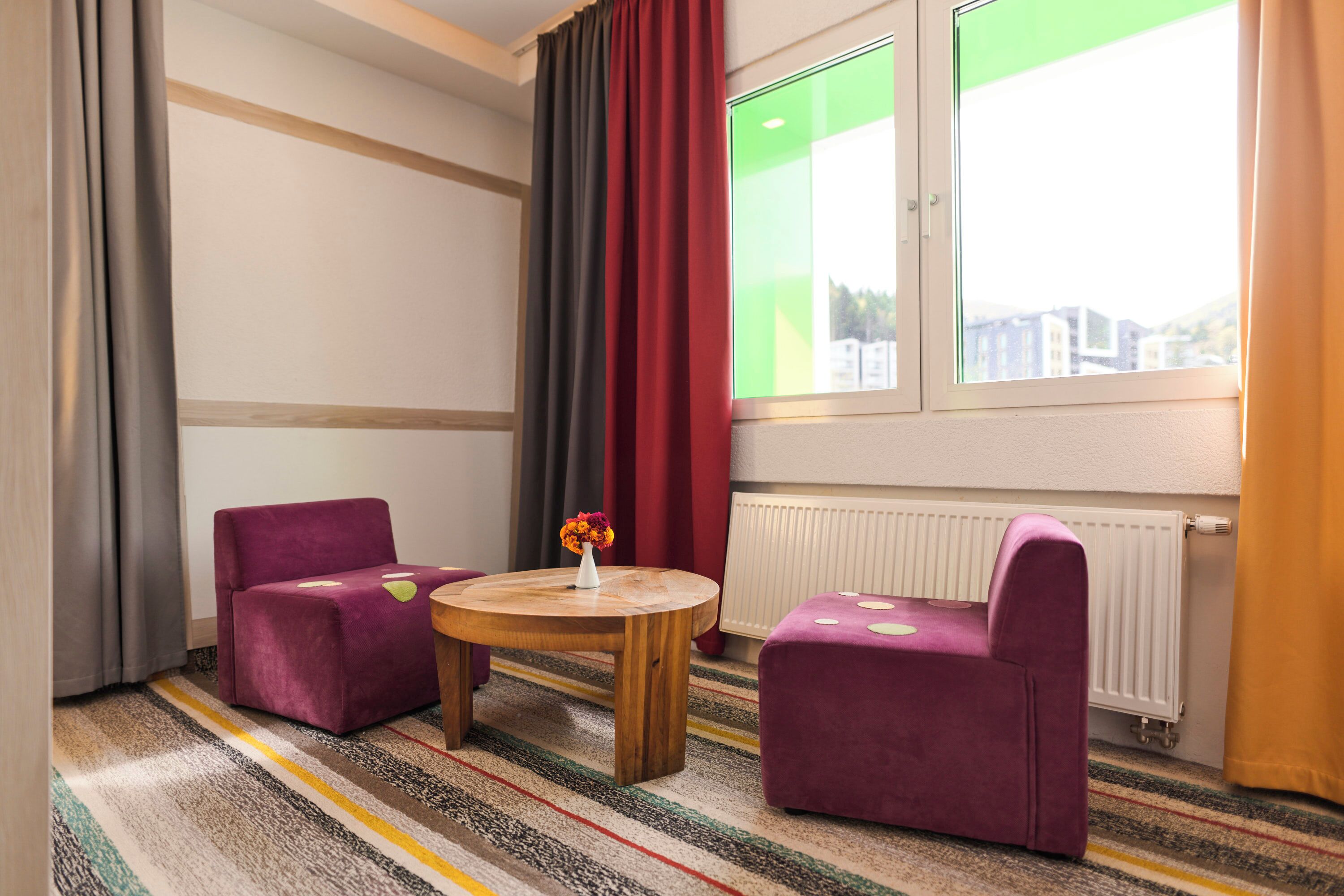 Una habitación de hotel con alfombra a rayas, dos sillones morados, una mesa de madera con un jarrón de flores y una ventana con vistas a un paisaje verde.