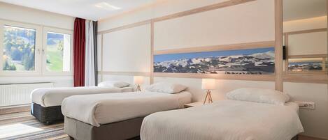 A hotel room with two beds, a large window with a view of greenery, a painting of mountains, and a striped carpet.