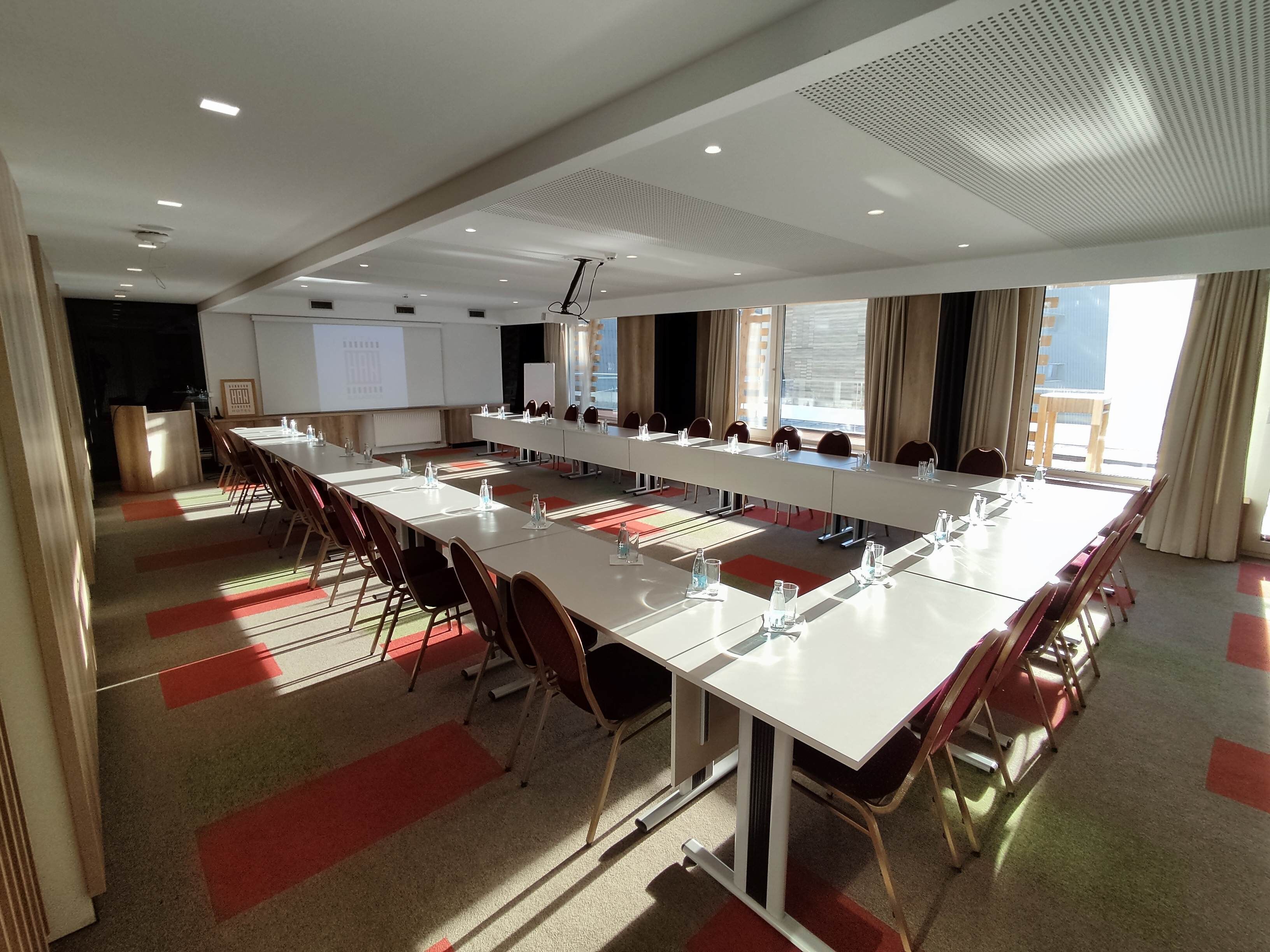Een vergaderzaal met lange tafels, stoelen en een projectorscherm.