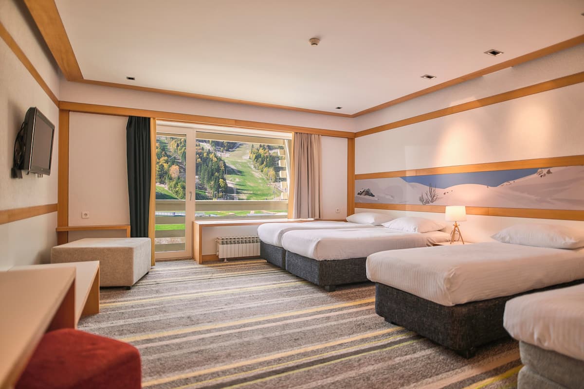 A hotel room with two beds, a large window with a view of a green landscape, a television on the wall, and a red chair.