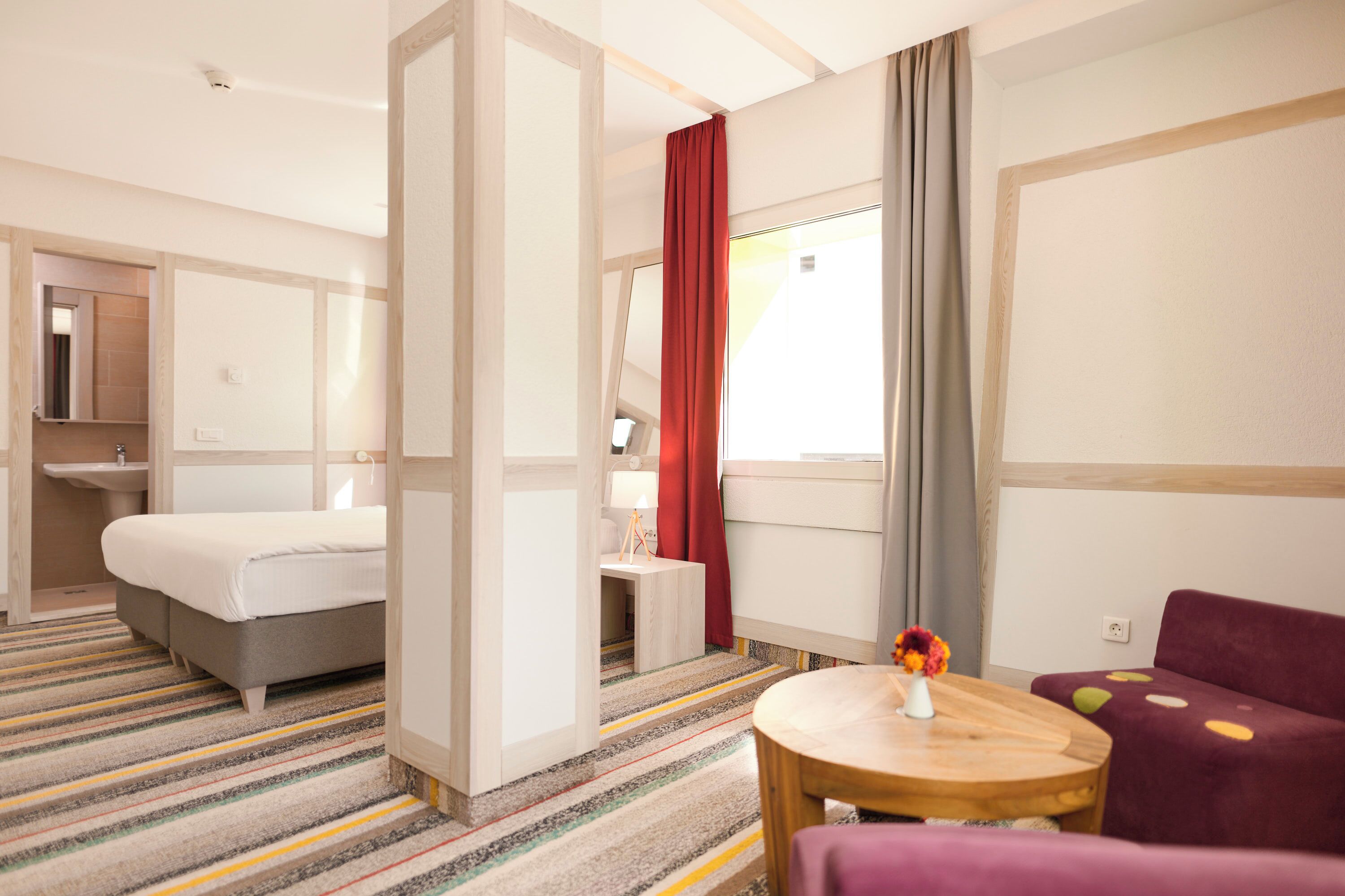 A hotel room with a bed, a desk, a chair, a small table, and a window with curtains.