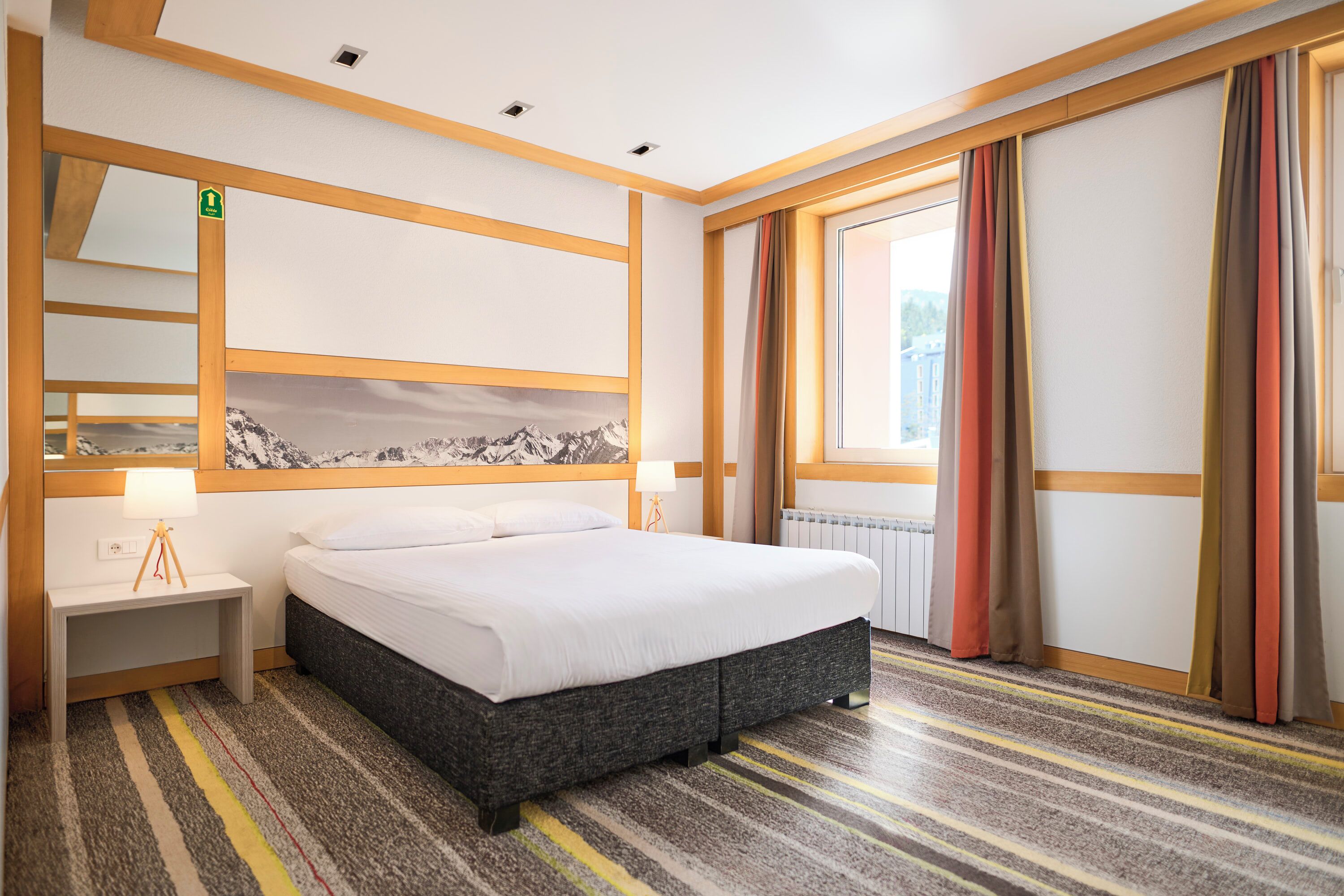 Une chambre d’hôtel avec un lit, une table de chevet, une lampe, une fenêtre avec des rideaux et une vue sur les montagnes.