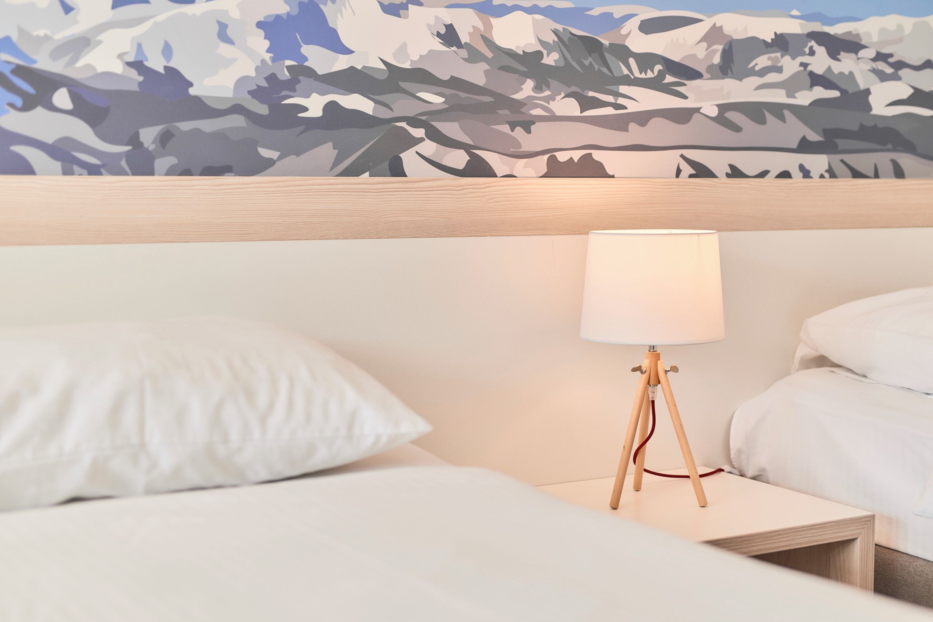 Een hotelkamer met een bed, nachtkastje, lamp en een muurschildering van een berglandschap.