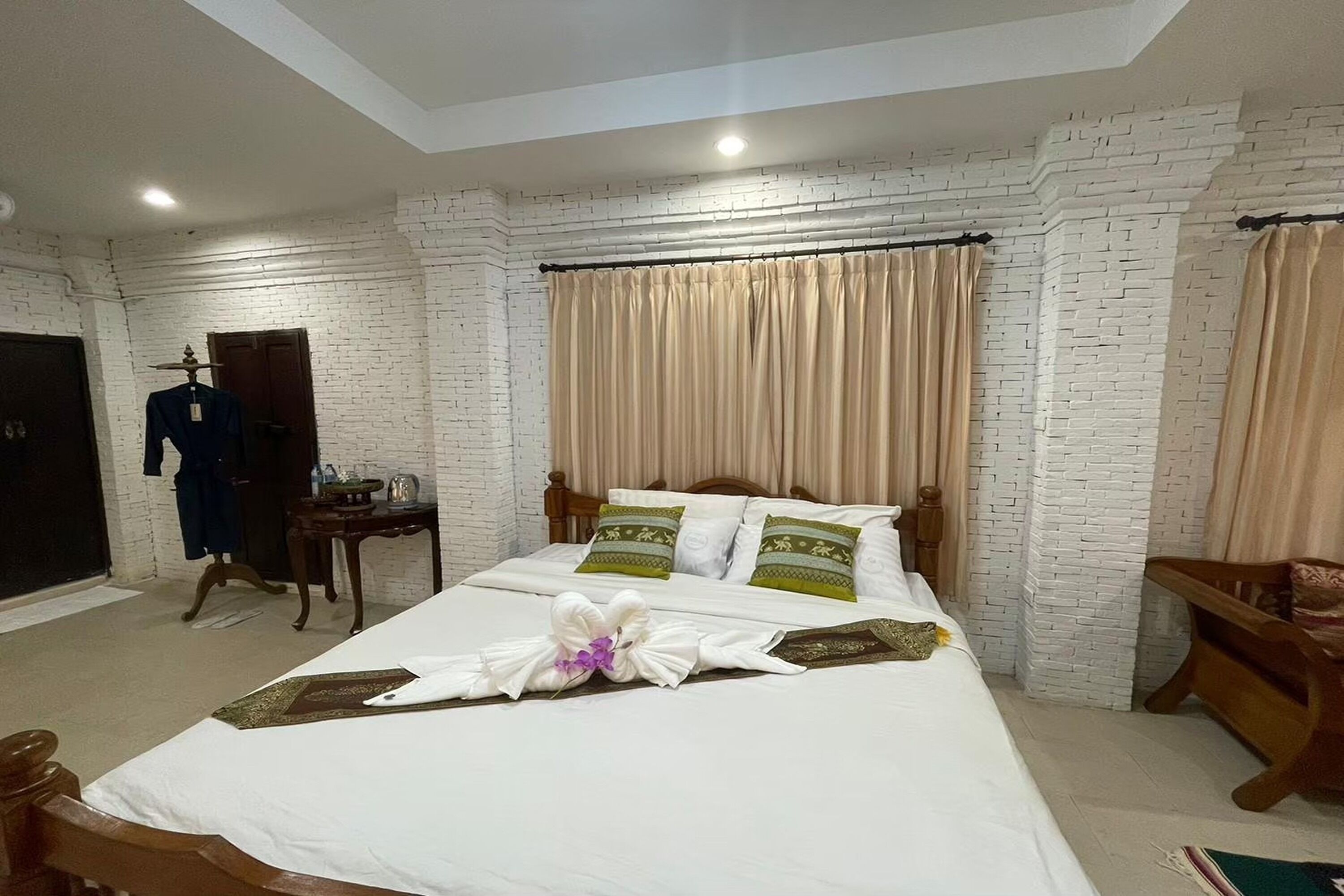 Habitación de hotel con pared de ladrillo blanco, cama con almohadas verdes y blancas, mesita de noche de madera y una silla de madera.