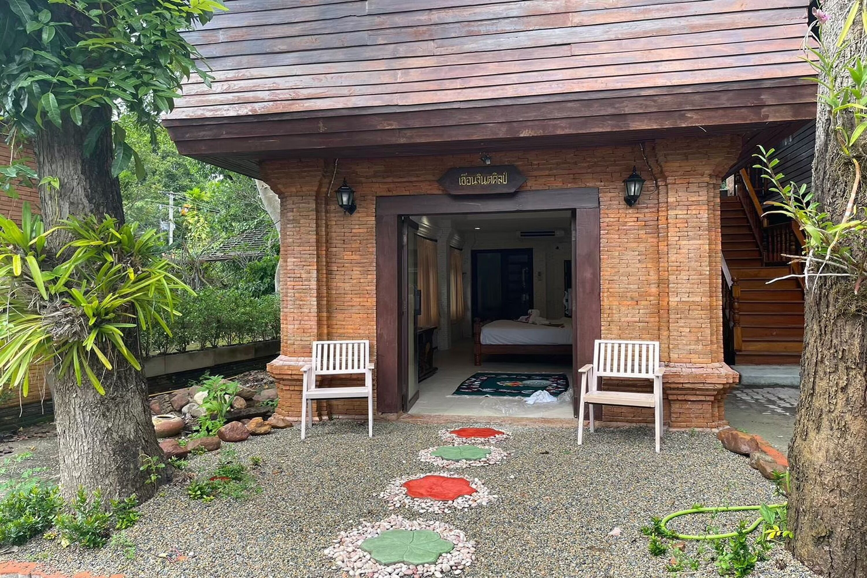 A rustic building entrance with a wooden overhang, two white chairs outside, and a gravel pathway with colorful stepping stones.
