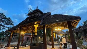 A traditional wooden pavilion with a thatched roof, surrounded by a garden with potted plants and a stone bench.
