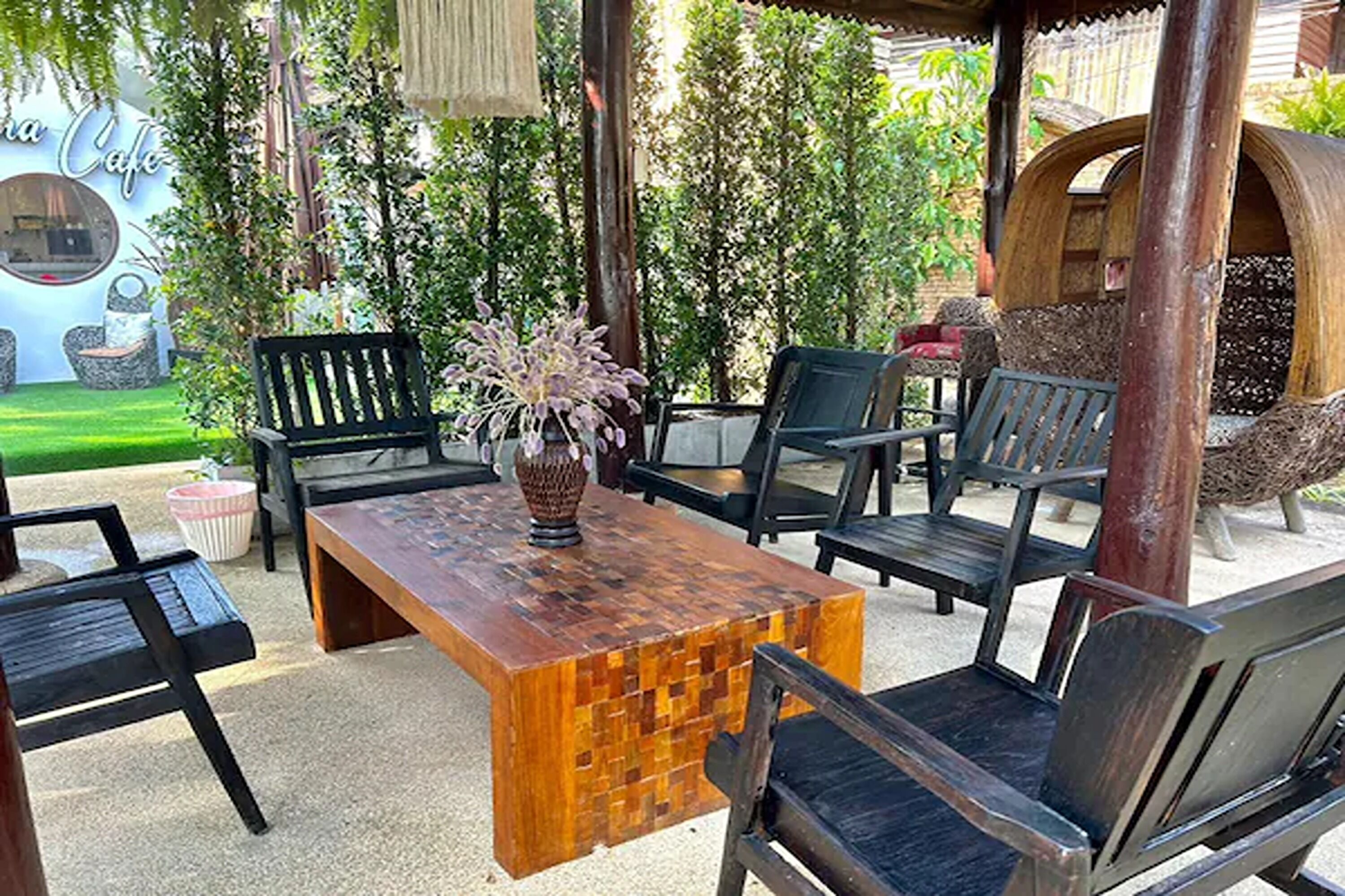 Un espacio al aire libre con mesa de madera y sillas negras, rodeado de follaje y una hamaca.